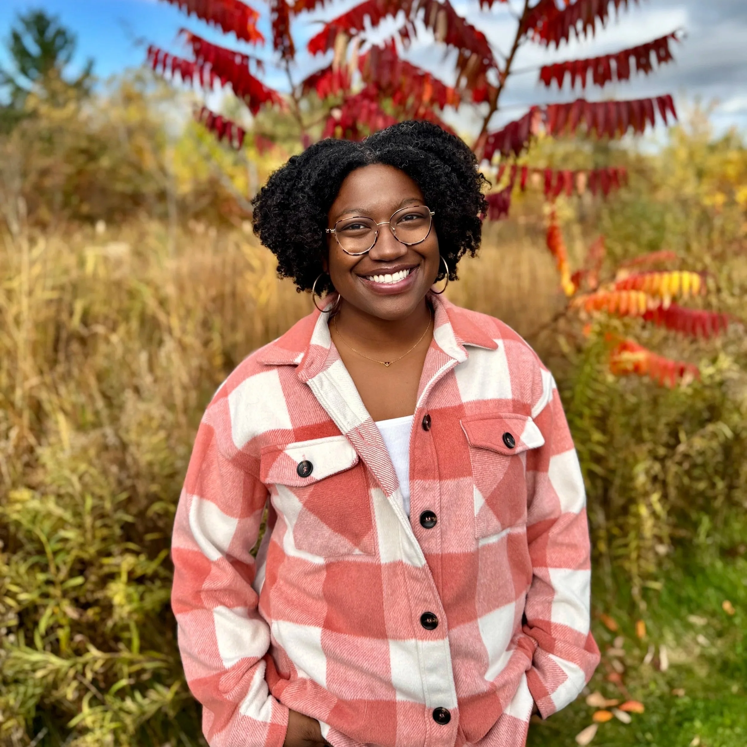 A young woman with curly hair and glasses smiling outdoors in fall, wearing a pink and white checkered jacket, with colorful autumn foliage in the background.