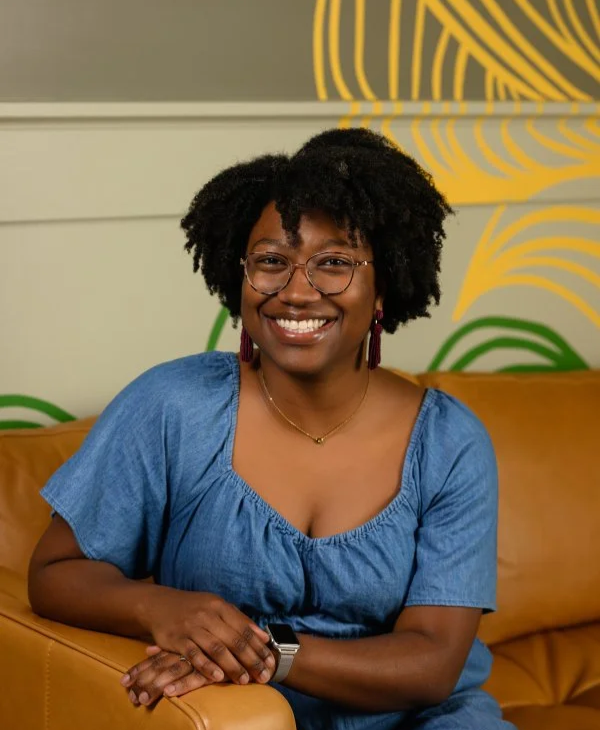 A woman with natural curly hair, glasses, and earrings smiling and sitting on a tan couch in front of a colorful abstract wall art.