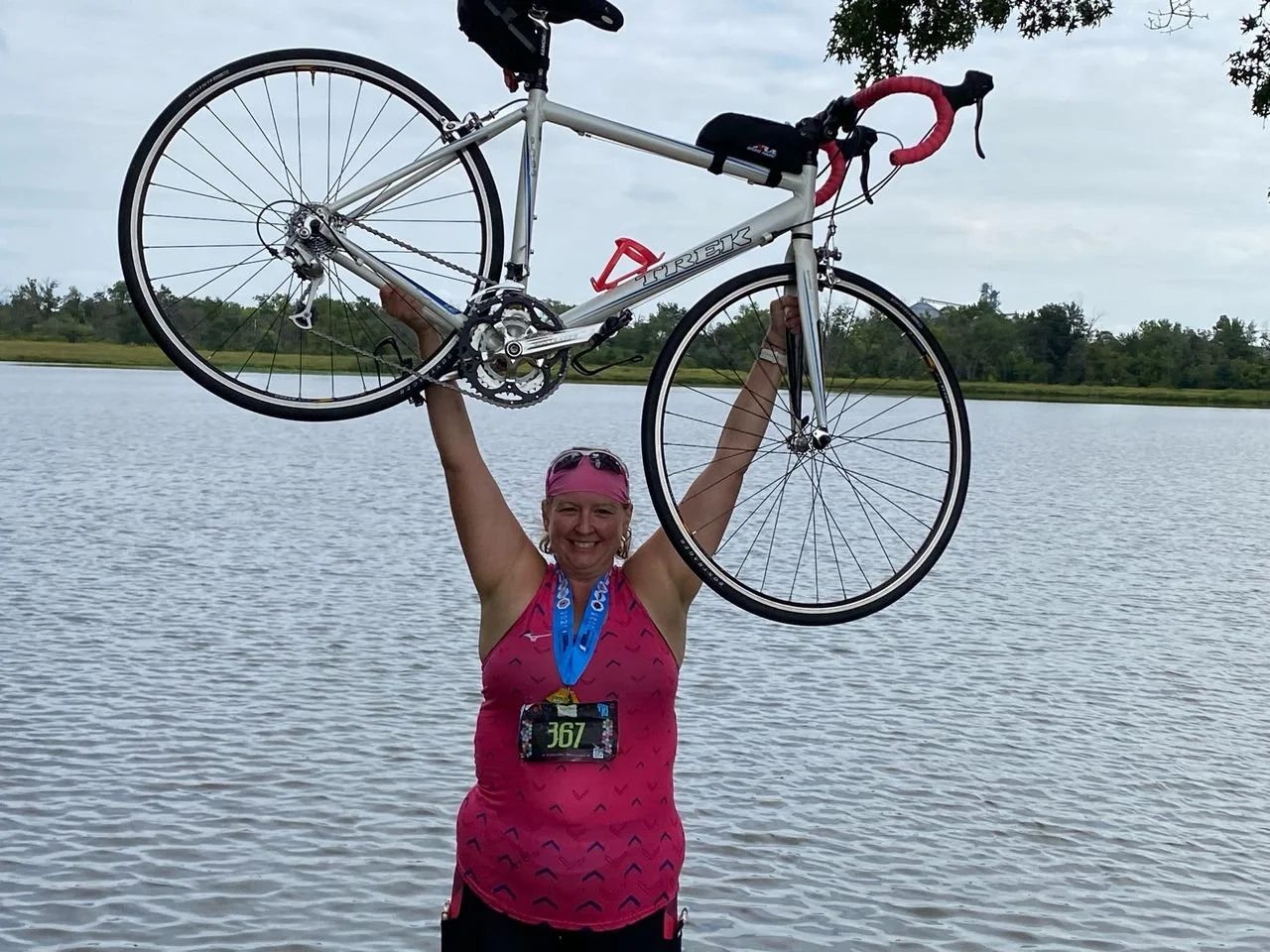 A woman in athletic gear holding a bicycle overhead near a body of water, smiling at the camera.