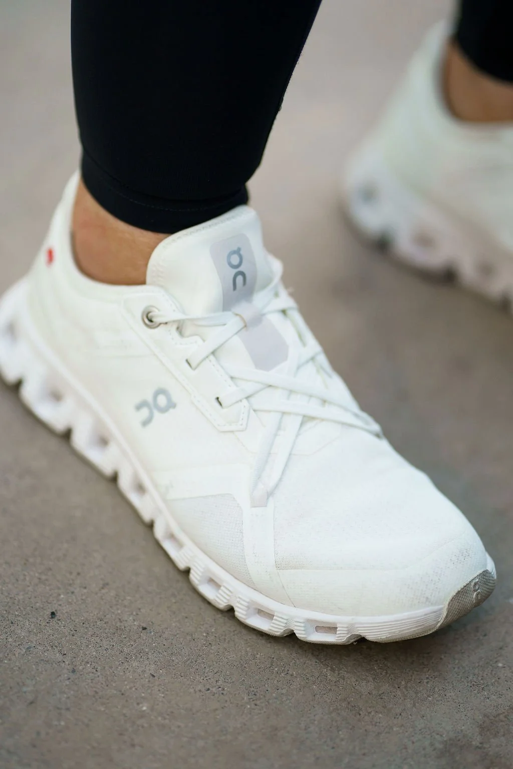 Person wearing white athletic sneakers with gray logos, black leggings, and standing on a gray surface.