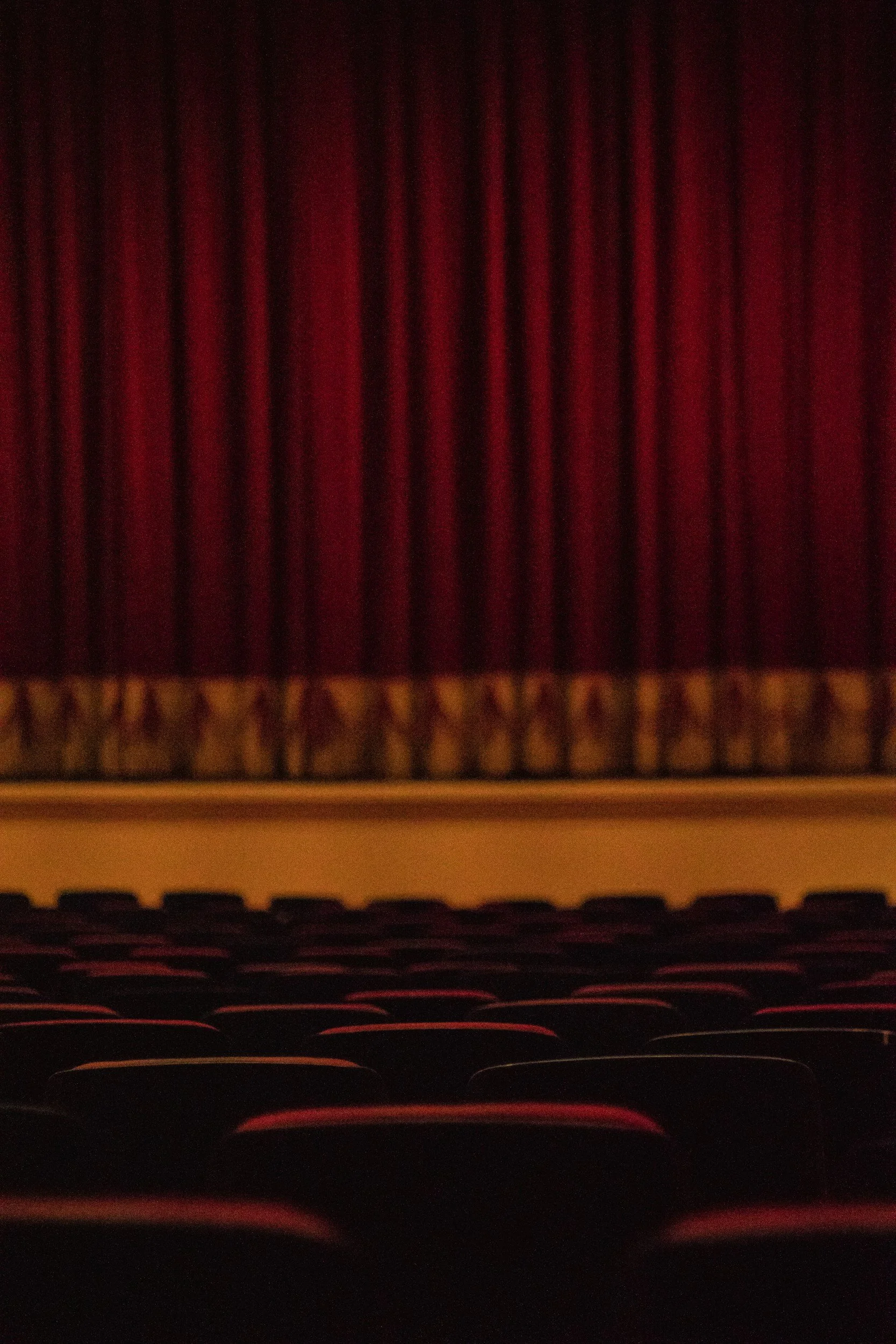 Inside of a theater with a view of the closed red velvet curtain and empty seats.