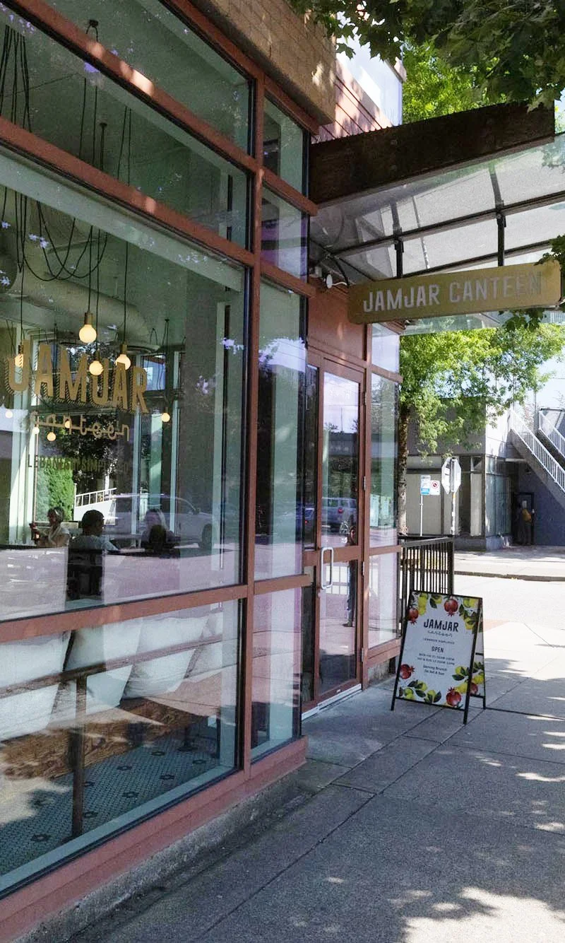 Exterior view of Jamjar Canteen showing glass windows, a door, and a sidewalk chalkboard sign outside with colorful fruit illustrations, indicating the restaurant is open.