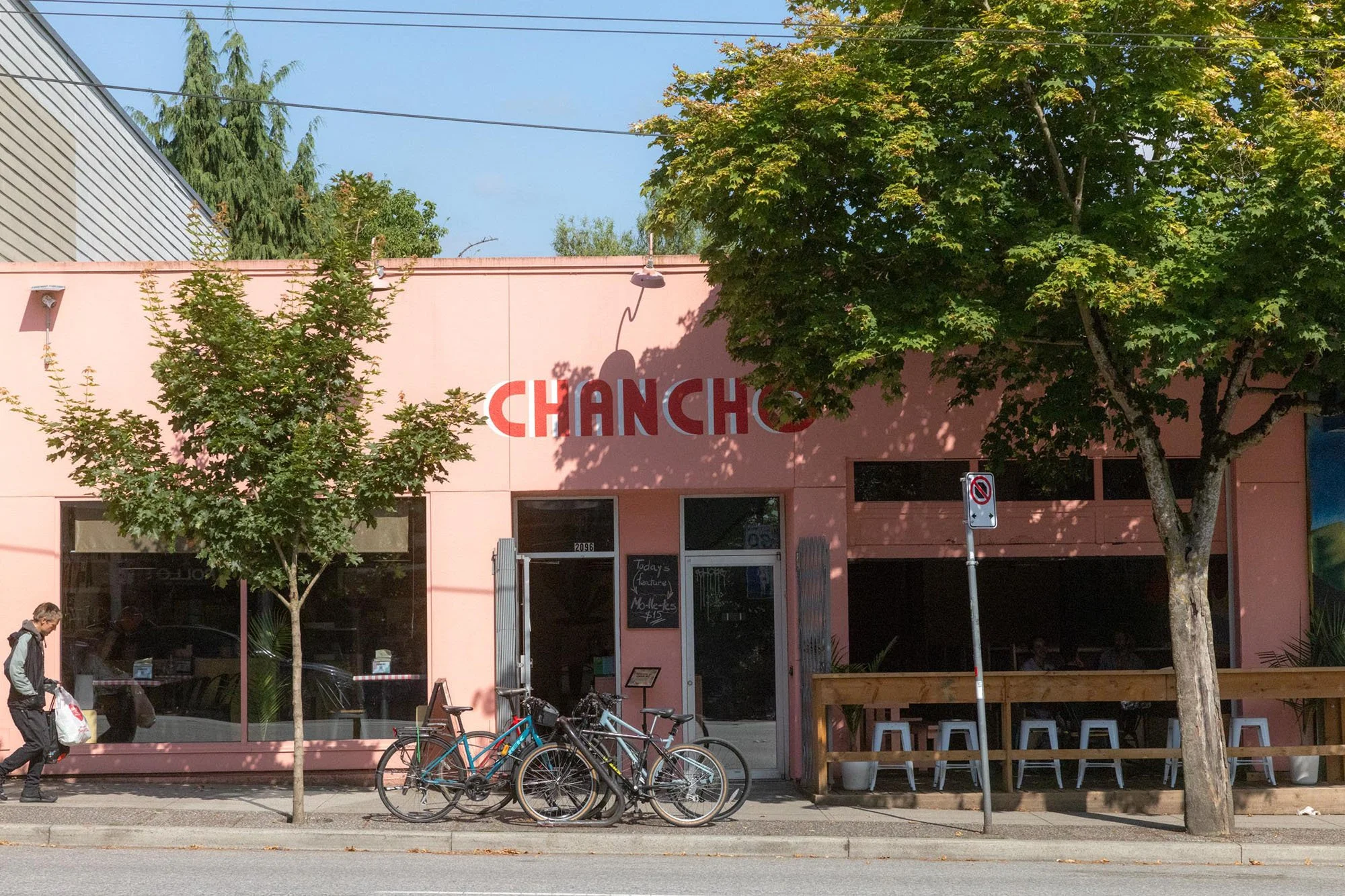 Pink storefront with red and white signage that reads 'CHANCH'. Front window displays with various items inside, exterior seating area with stools, a no parking sign, bicycles parked outside, and trees providing shade on the sidewalk.