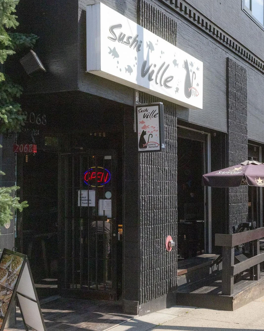 Exterior of a Japanese restaurant named Sushi Ville with a sign featuring a black cat and fish. The restaurant is open, indicated by a red 'OPEN' sign in the window, and has outdoor seating with umbrellas.