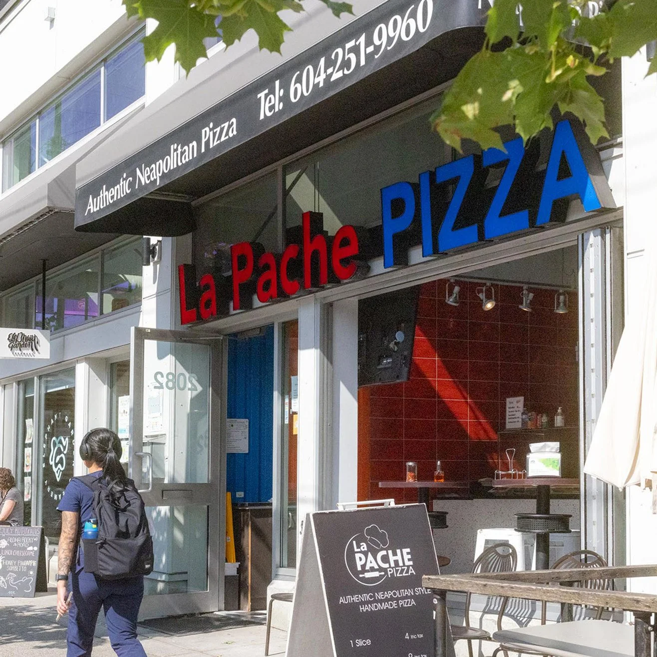 Exterior view of La Pache Pizza restaurant with a black and red sign, open door, sidewalk seating, and a woman walking by wearing a backpack.