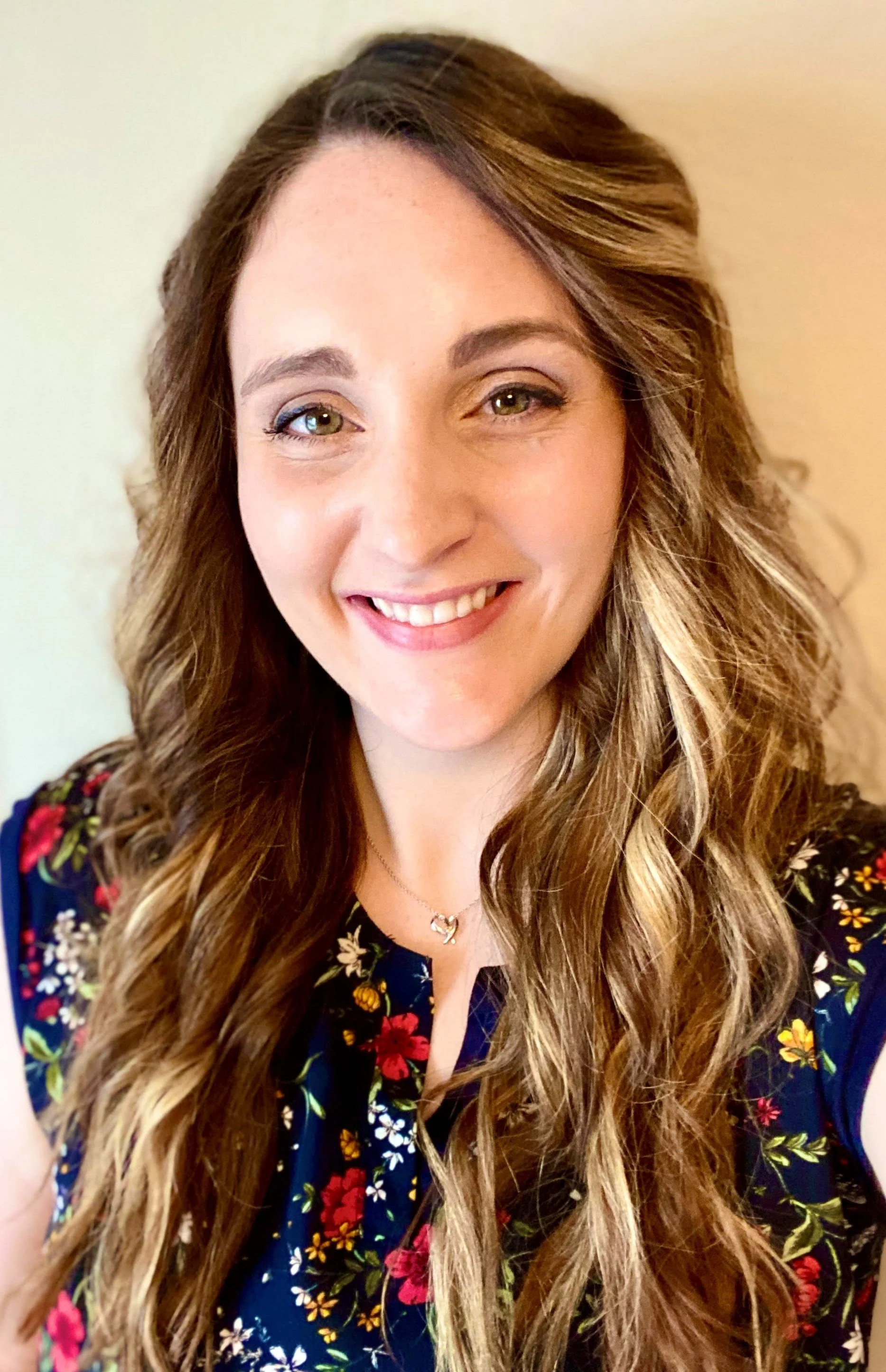 A woman with long, wavy light brown hair and green eyes, smiling, wearing a floral blouse and a necklace with a heart-shaped pendant.
