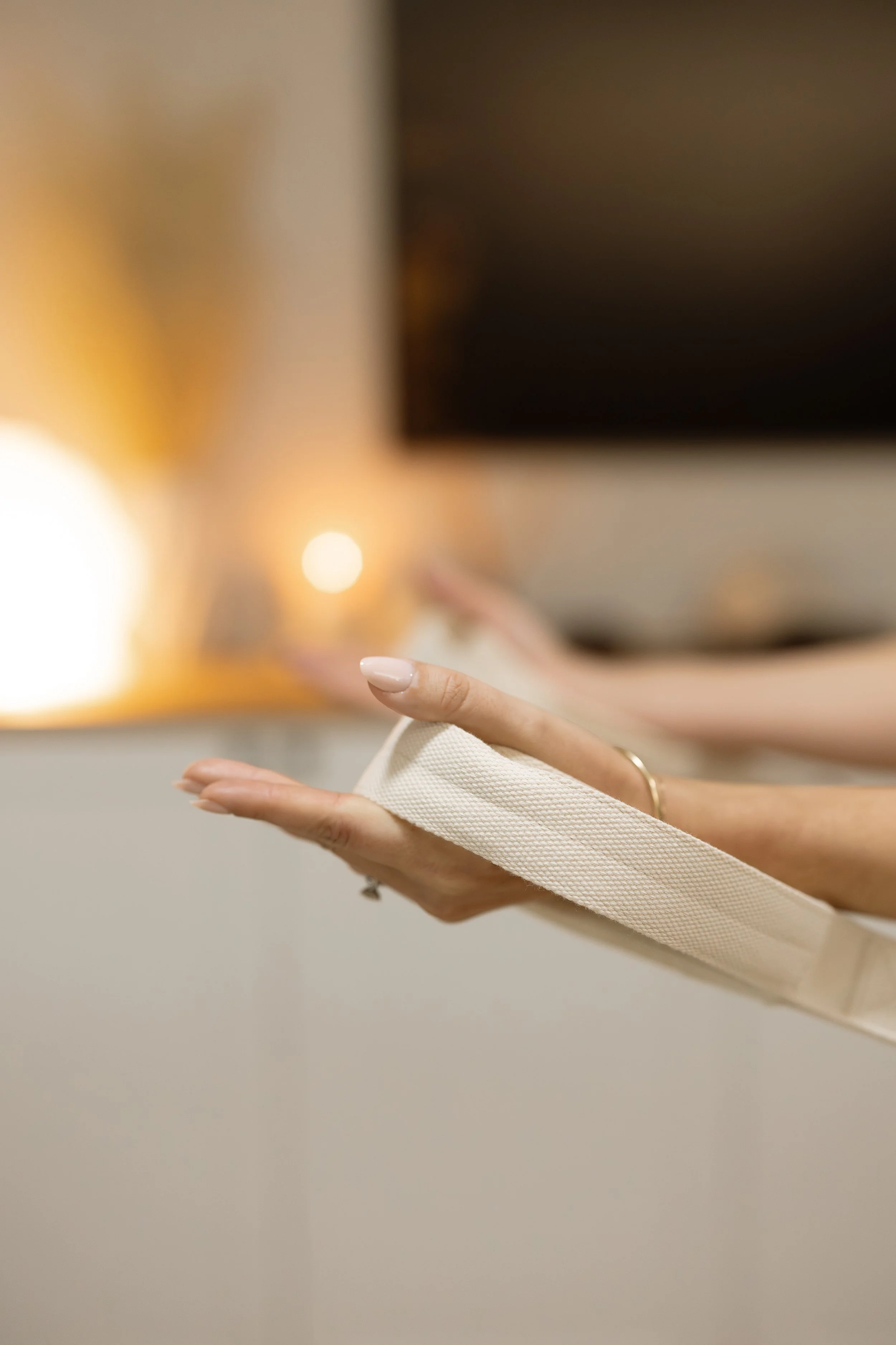 Person holding a beige canvas strap of a reformer pilates machine with one hand, in an at home pilates studio with warm lighting.
