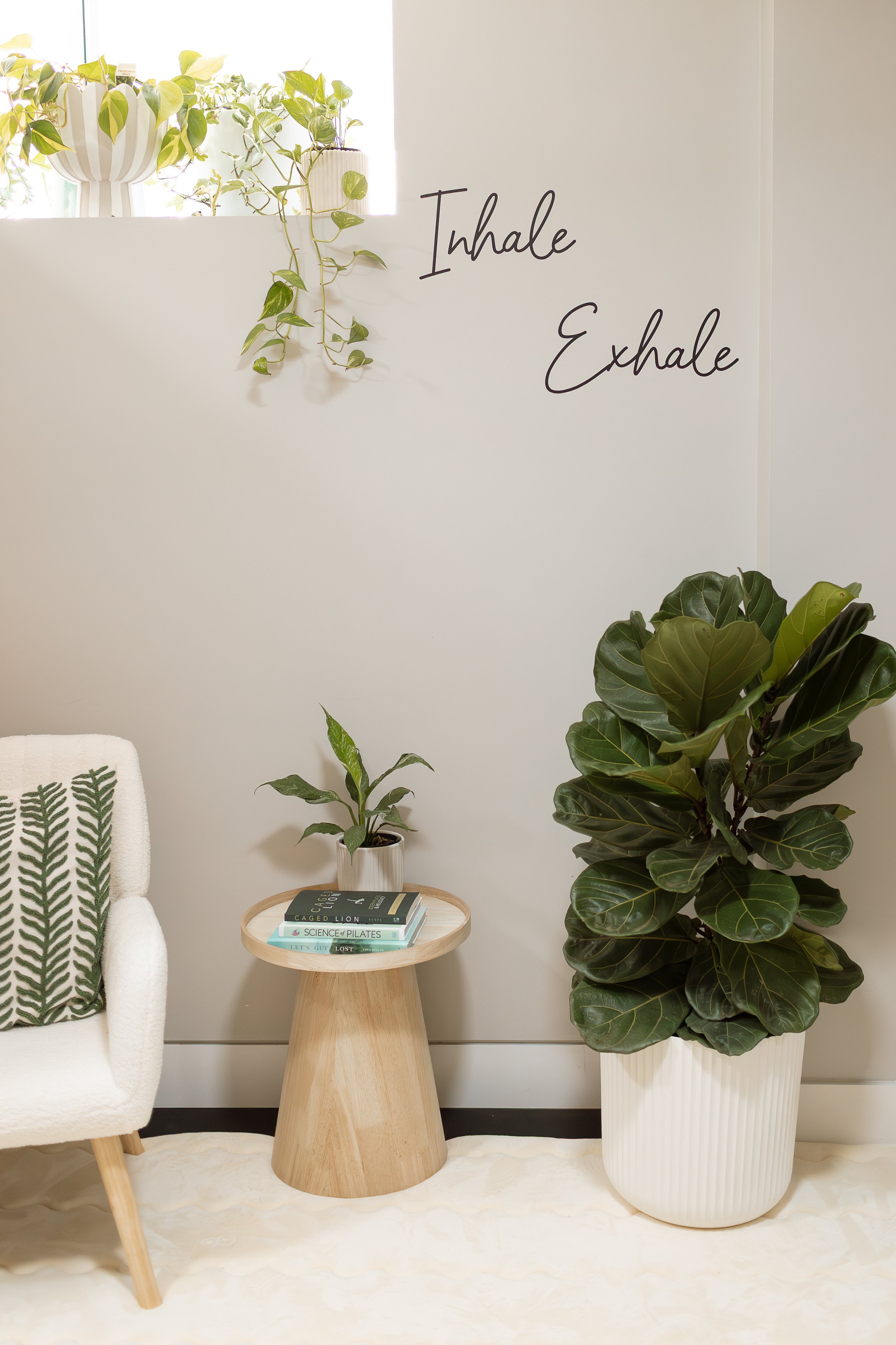 A cozy corner with a white armchair with a green leaf patterned pillow, a small round wooden side table holding books and a plant, and a large potted fiddle leaf fig plant. There are wall decals saying 'Inhale' and 'Exhale' and a corner shelf with potted plants and a window with sunlight.