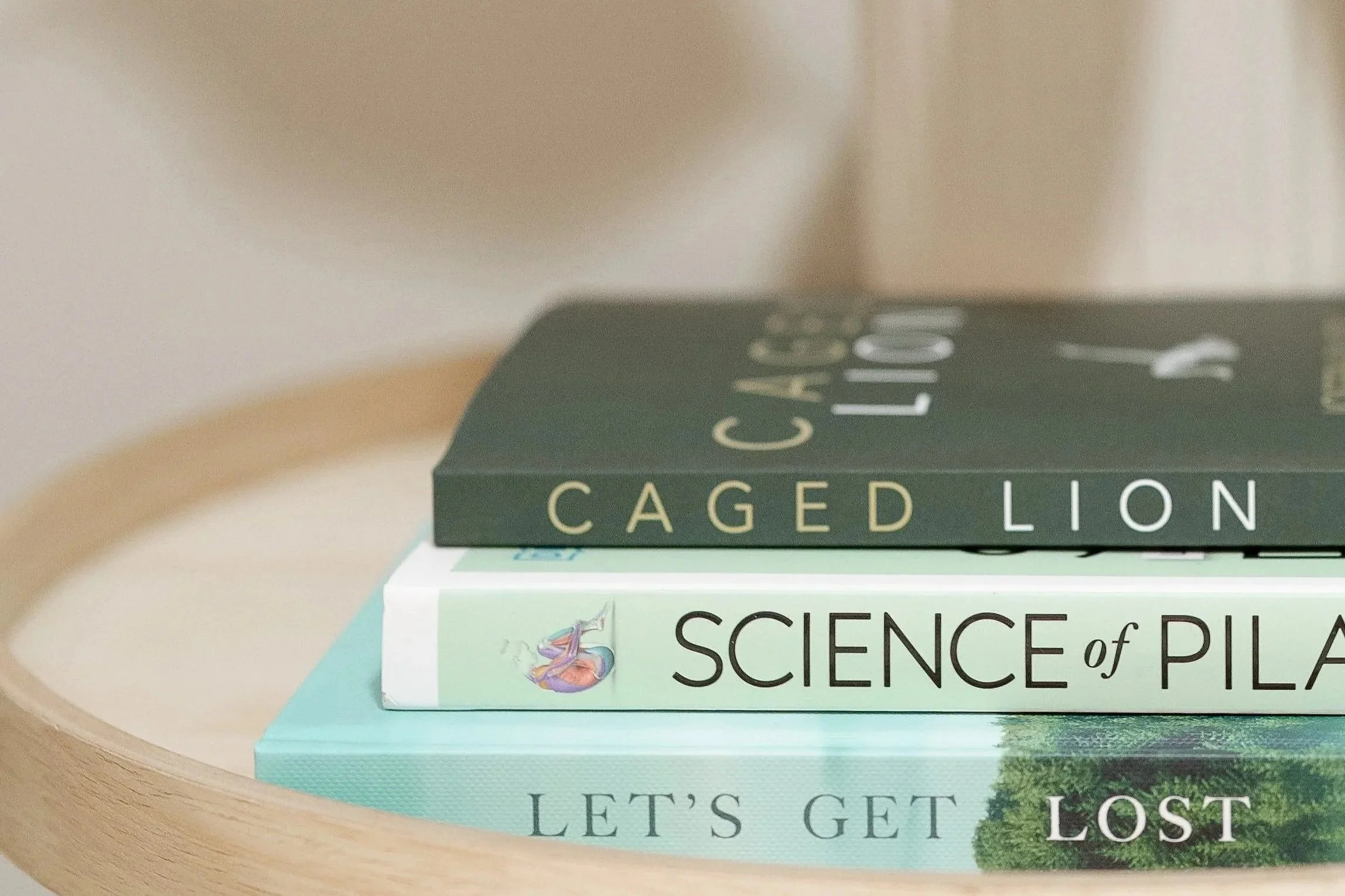 Close-up of three stacked books on a wooden surface, with titles "Caged Lion," "Science of Pilates," and "Let's Get Lost."