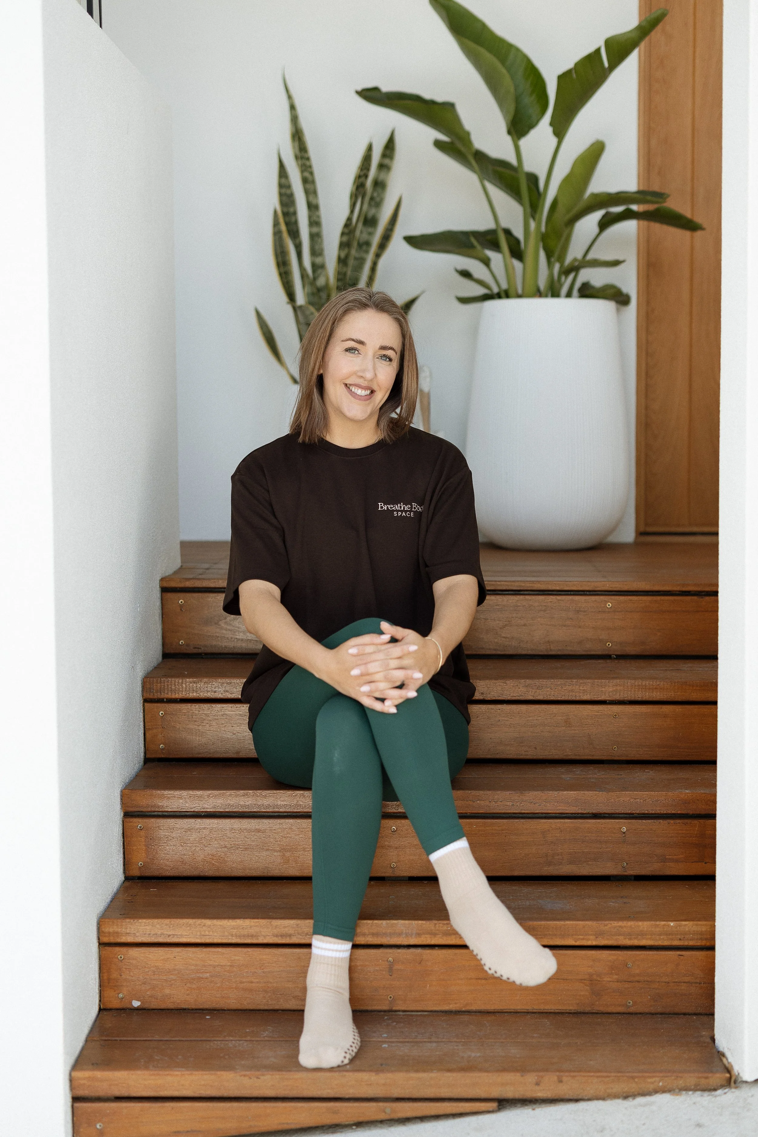 A pilates teacher sitting on the front steps of the entrance to an at home pilates studio, smiling, wearing a brown t-shirt, green leggings, and beige socks with white details, with large potted plants in the background.