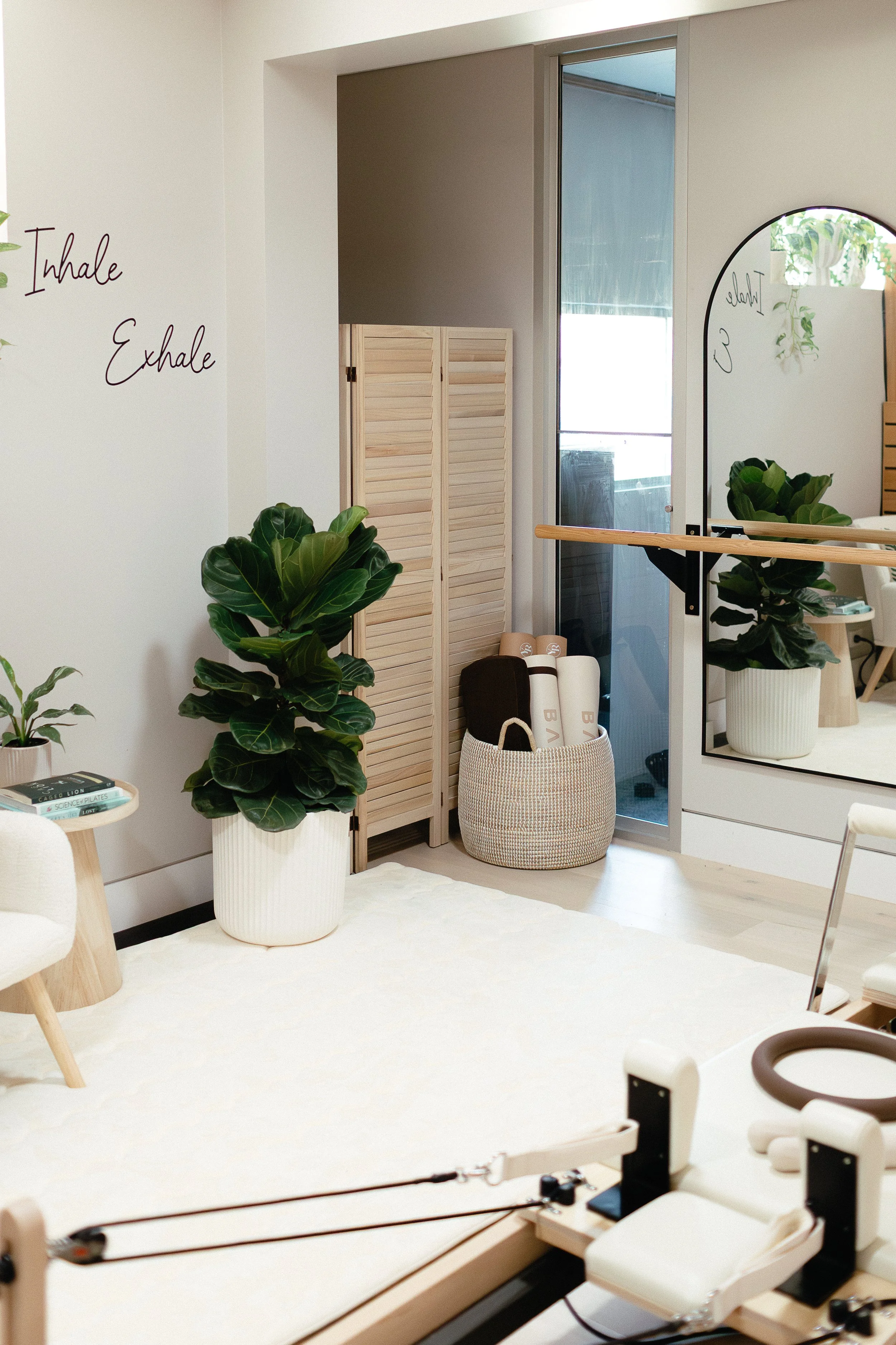 A pilates studio interior with a sign on the wall reading 'Inhale, Exhale', green plants in white pots, a mirror, wooden folding room divider, fitness equipment. 