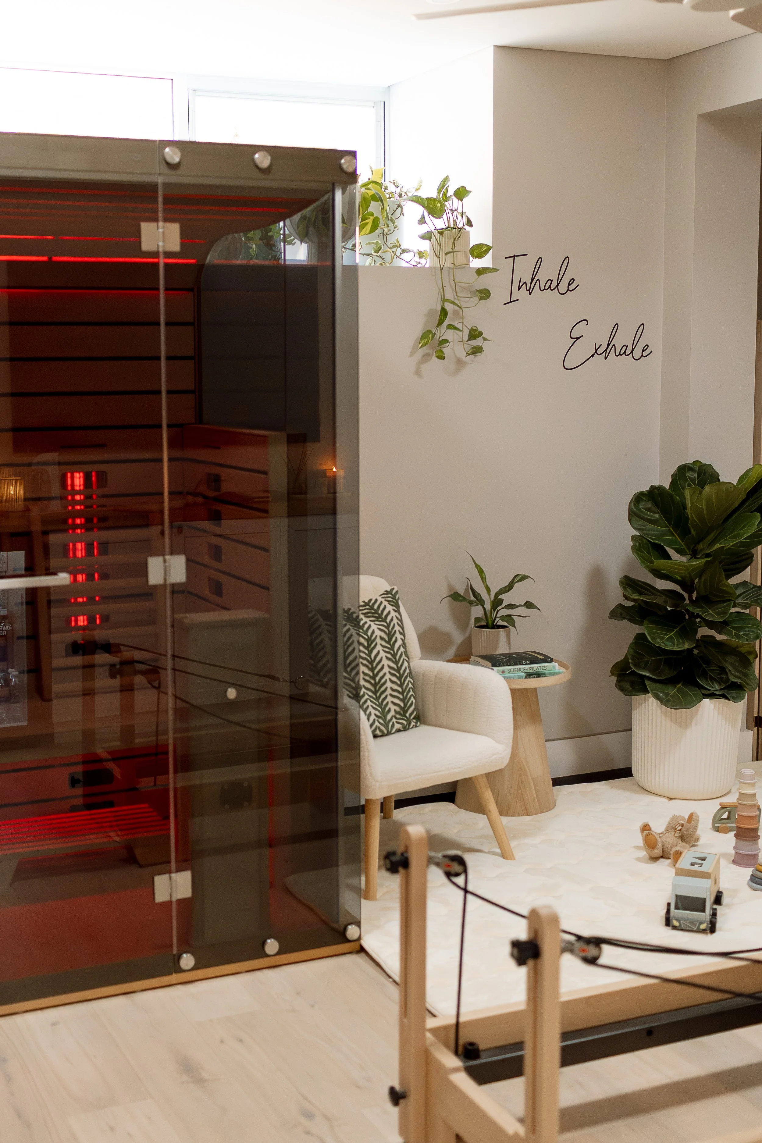 Pilates studio with an infrared sauna, a white armchair with patterned cushions, a small side table with potted plants, large potted fiddle leaf fig, and wall decor that reads "Inhale Exhale".