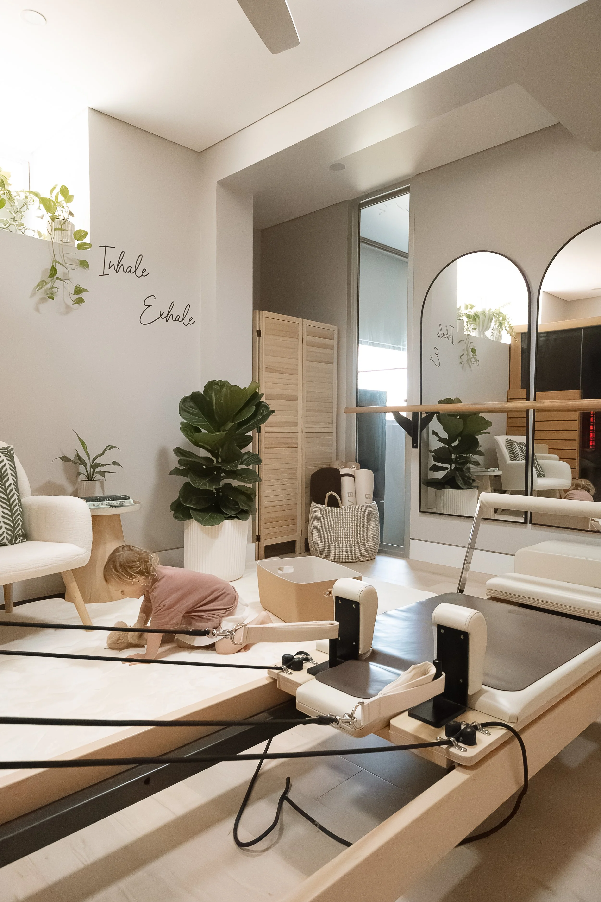 A young girl is kneeling on a cushioned surface in a modern, minimalistic at home pilates studio with white walls, plants, a large mirror, and a wooden room divider. The wall has the words 'Inhale' and 'Exhale' written on it.