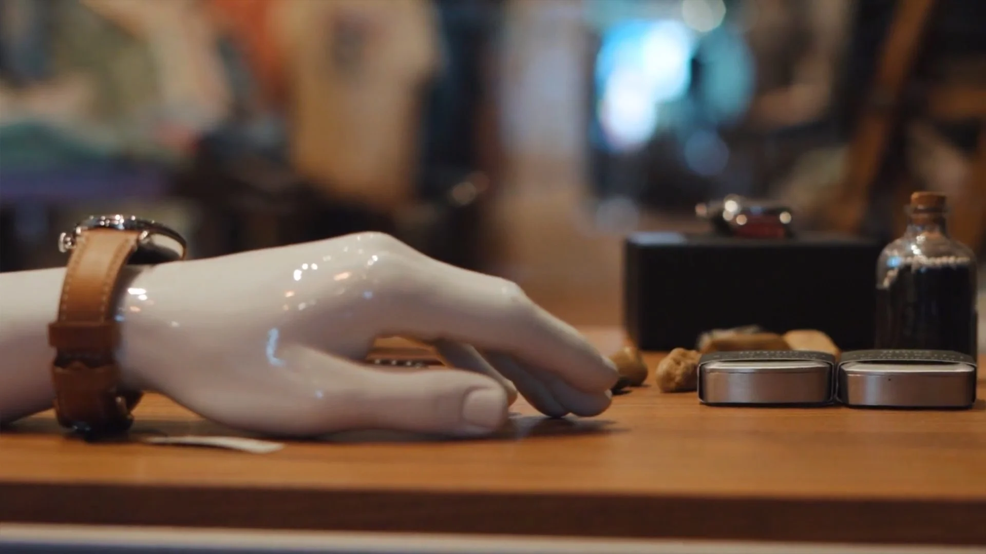 A porcelain hand mannequin with a brown leather watch resting on a wooden surface, with various small objects including tins, a small bottle, and rocks on the table, in a cozy, blurred background.