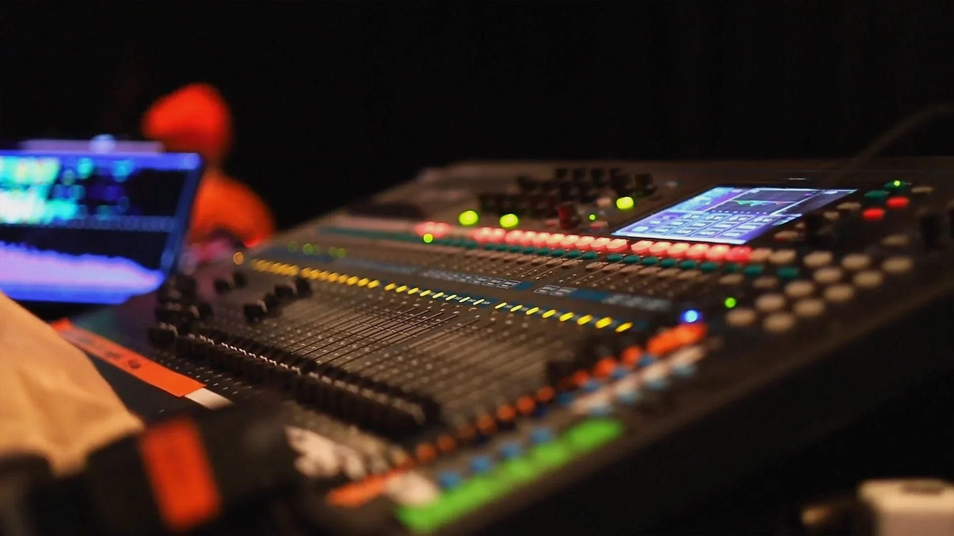 A close-up of a sound mixing console with various colored lights and sliders, focused on the equipment, with a person in a hoodie blurred in the background.