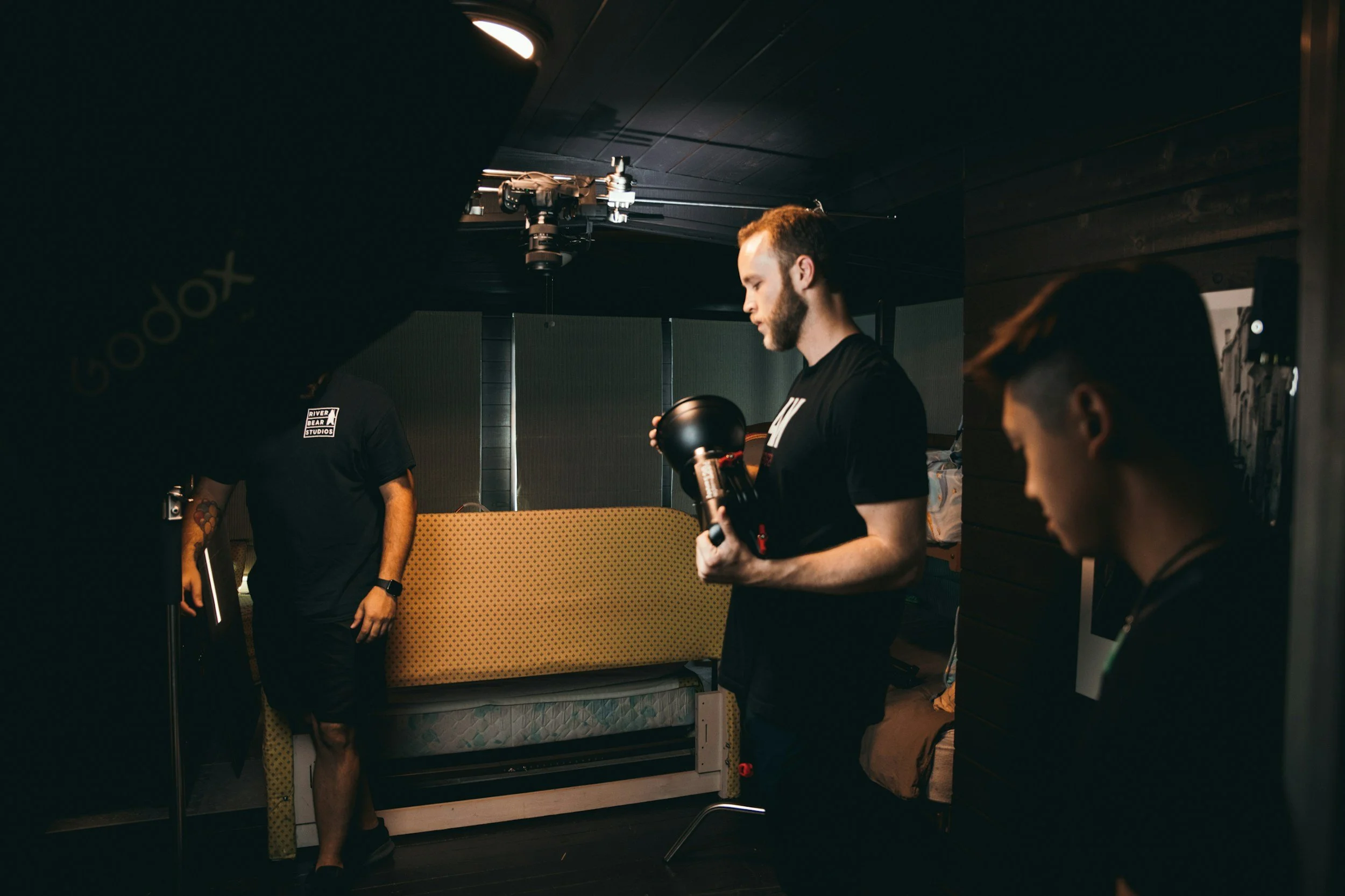 Three men in a dimly lit room, one holding a helmet, another with a monitor, and the third with a tablet, possibly preparing for a film or photo shoot.