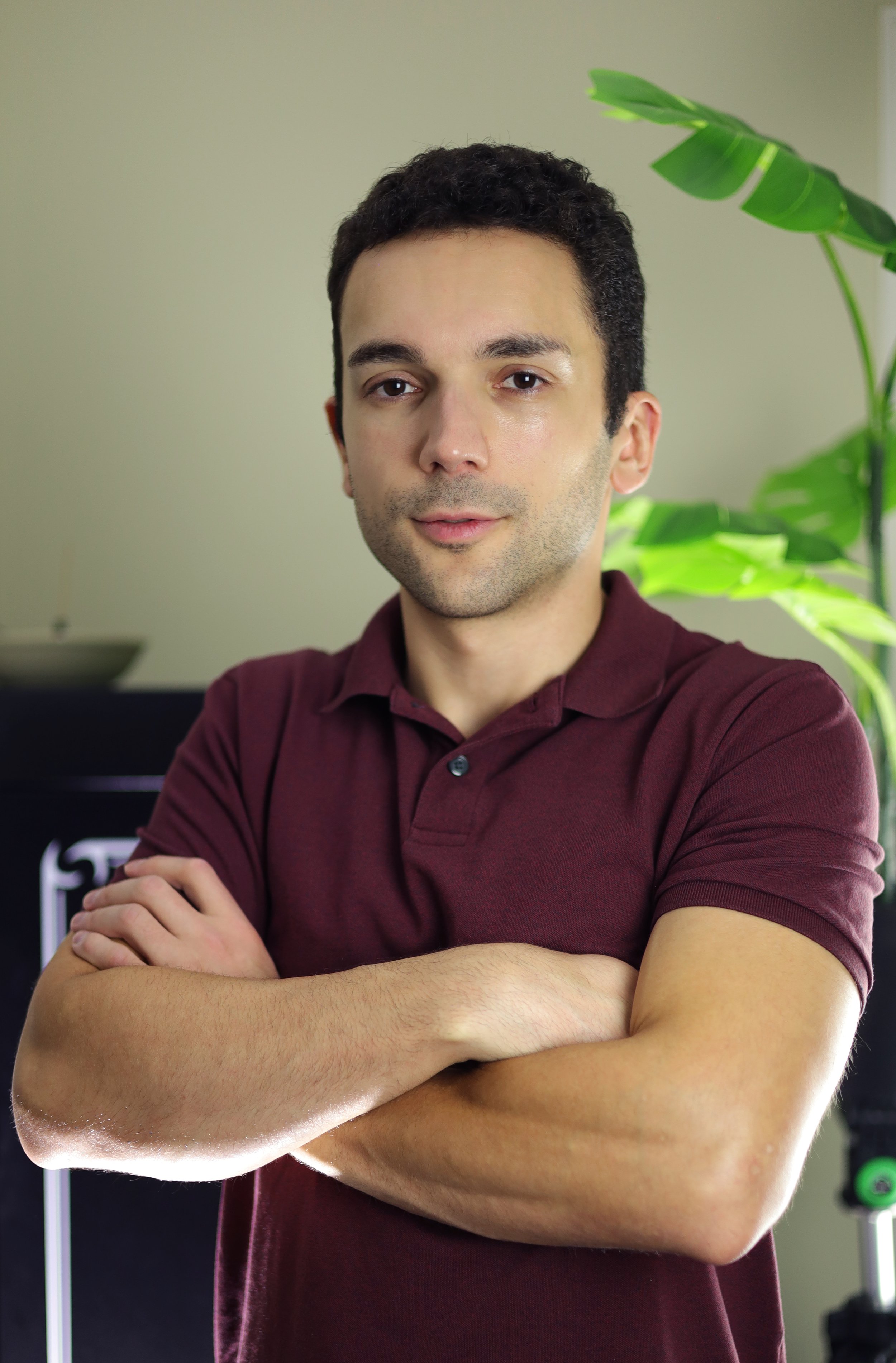 A man with short dark hair and light skin, wearing a burgundy polo shirt with arms crossed, standing indoors near a green plant.