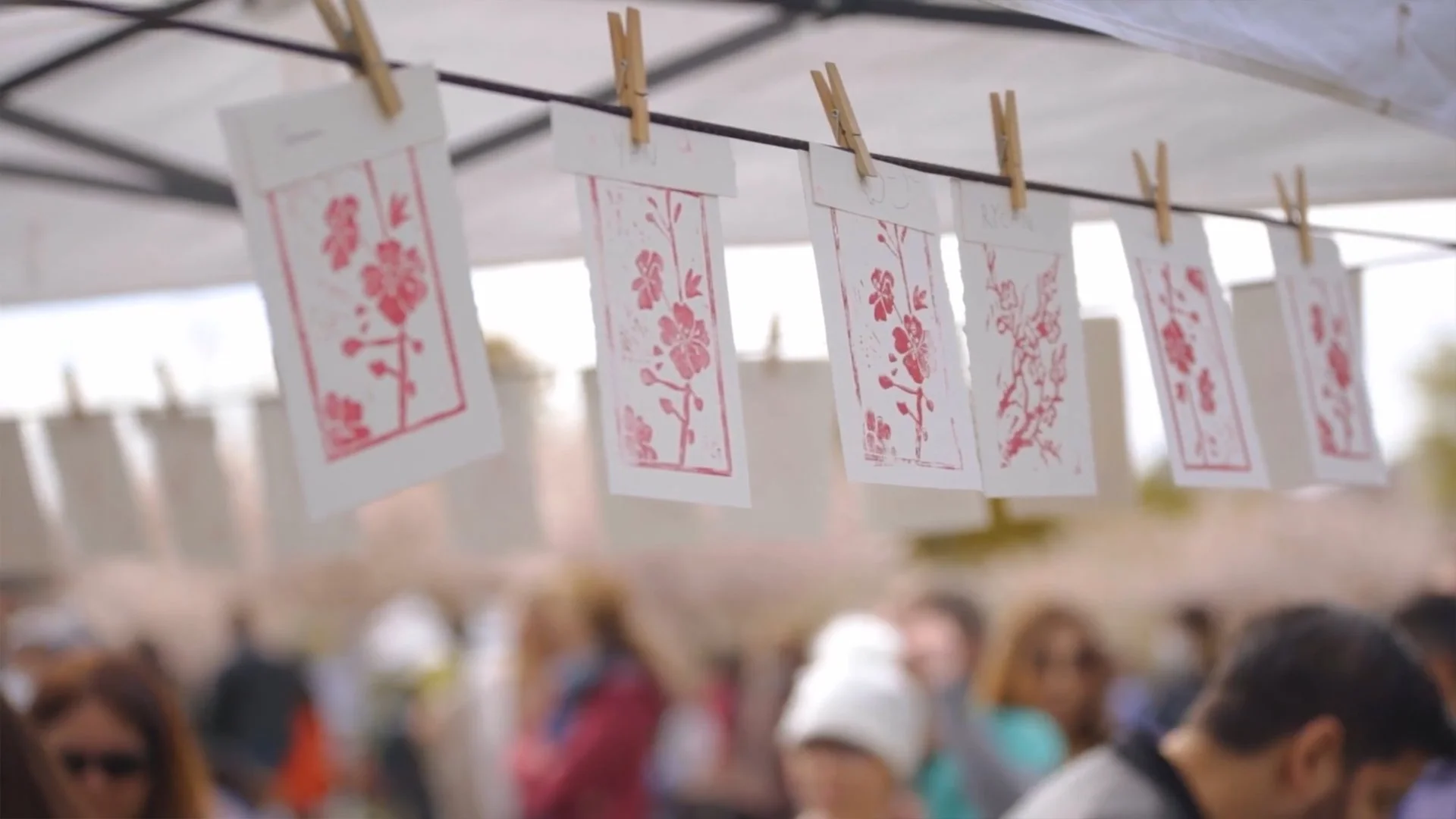 Paper artworks hanging on a string with clothespins at an outdoor event with a crowd of people in the background.