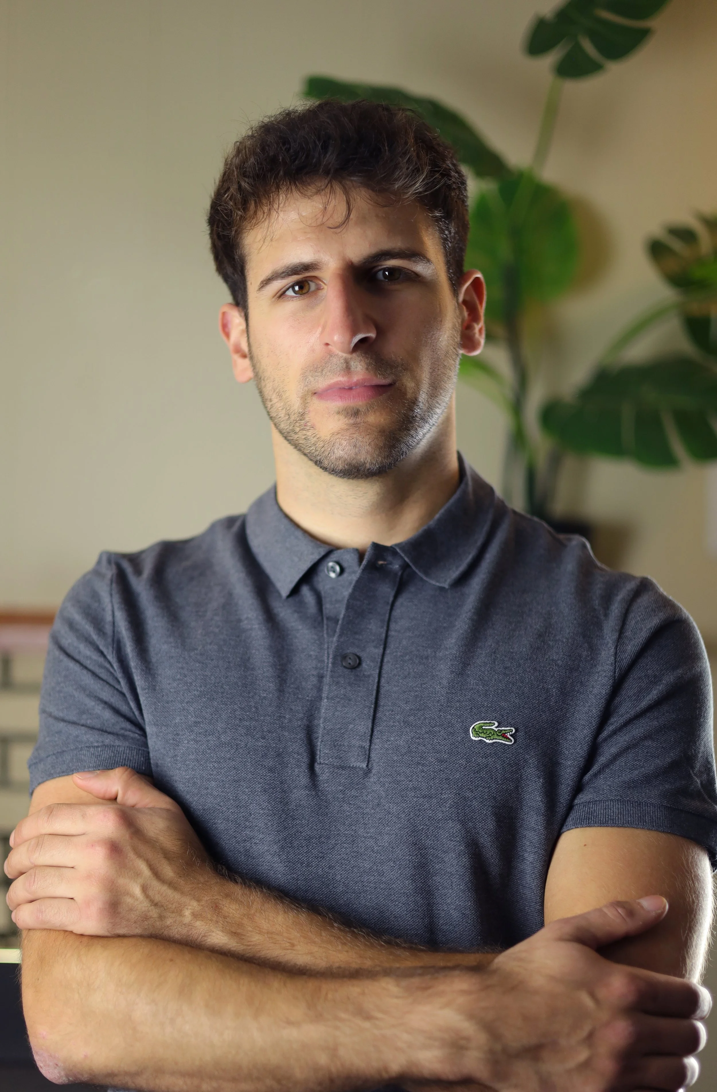 A man with short dark hair and a beard standing indoors, crossing his arms, wearing a gray polo shirt with a small green crocodile logo, with large green leafy plants in the background.