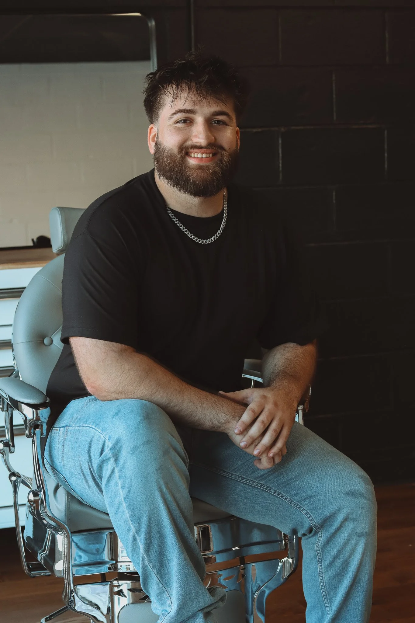 A man with a beard, wearing a black t-shirt, silver chain necklace, and light blue jeans, sitting on a chair in an indoor setting with a dark wall in the background, smiling at the camera.