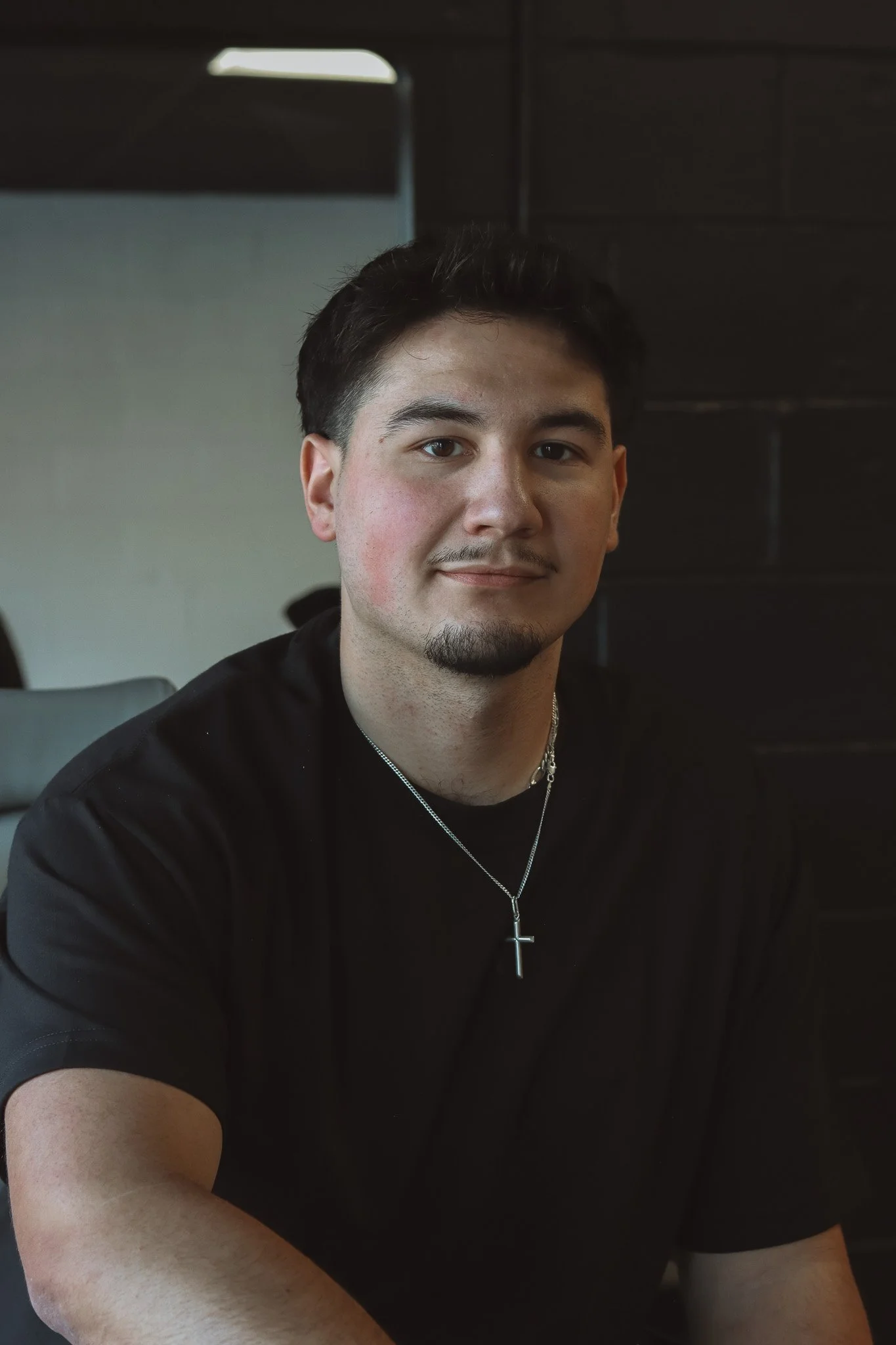 A young man with dark hair and a mustache, wearing a black shirt and a silver cross necklace, sitting indoors with a dark background.
