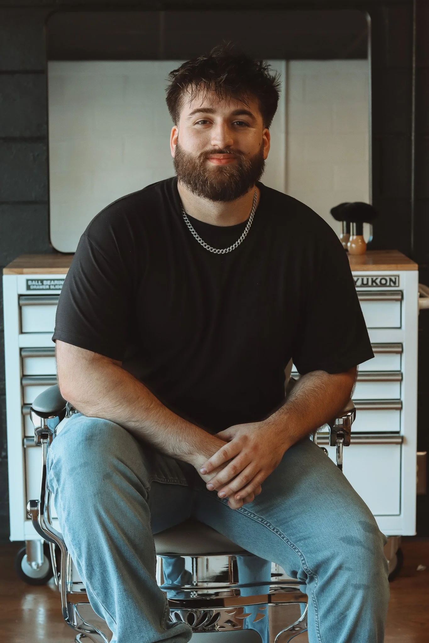 A young man with a beard and dark hair sitting in a modern office chair, wearing a black t-shirt and a silver chain necklace. Behind him is a white cabinet with multiple drawers and a large mirror. The setting appears to be a contemporary workspace or studio.
