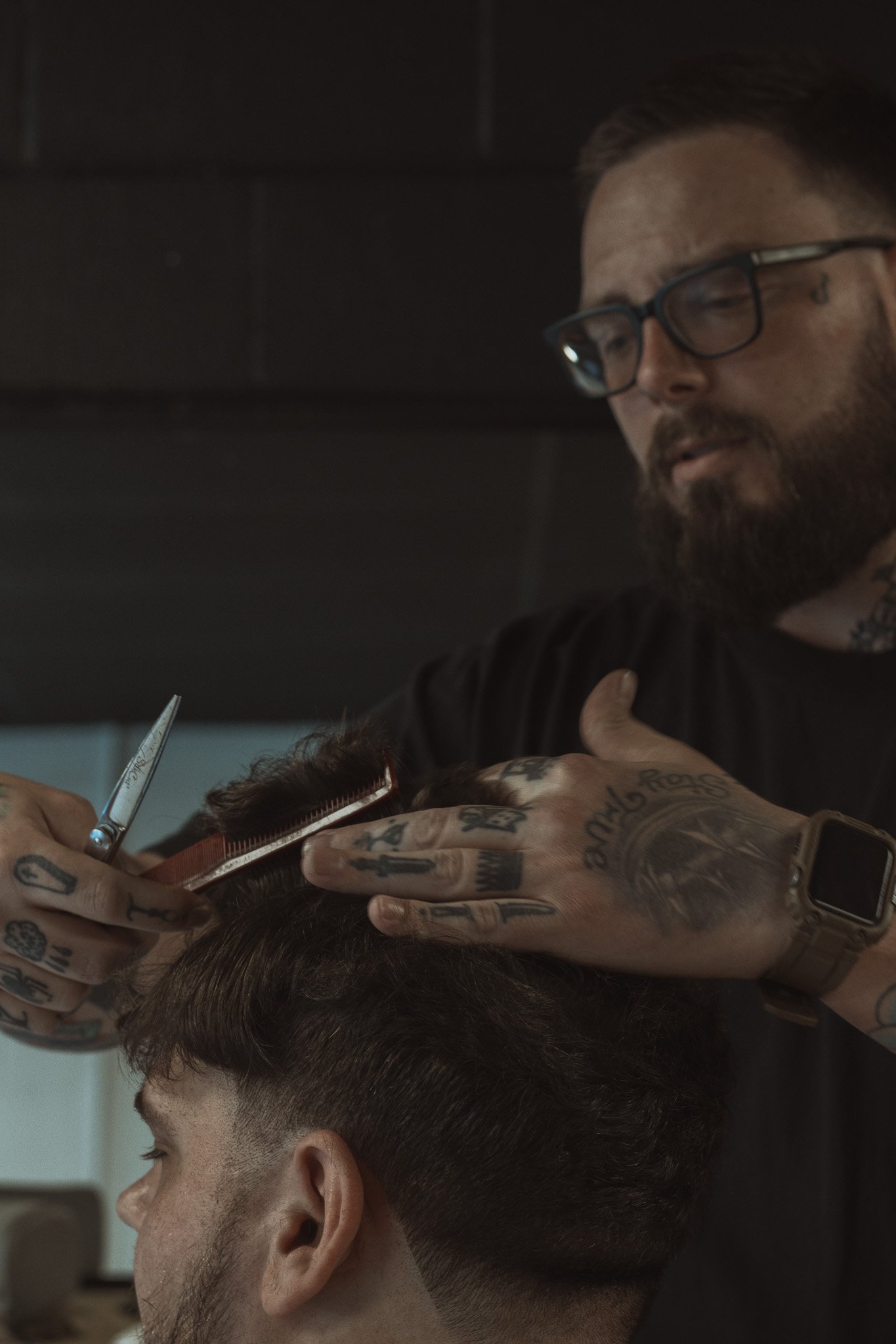 A tattooed barber with glasses and a watch is giving a haircut to a young man with dark hair, in a barbershop.