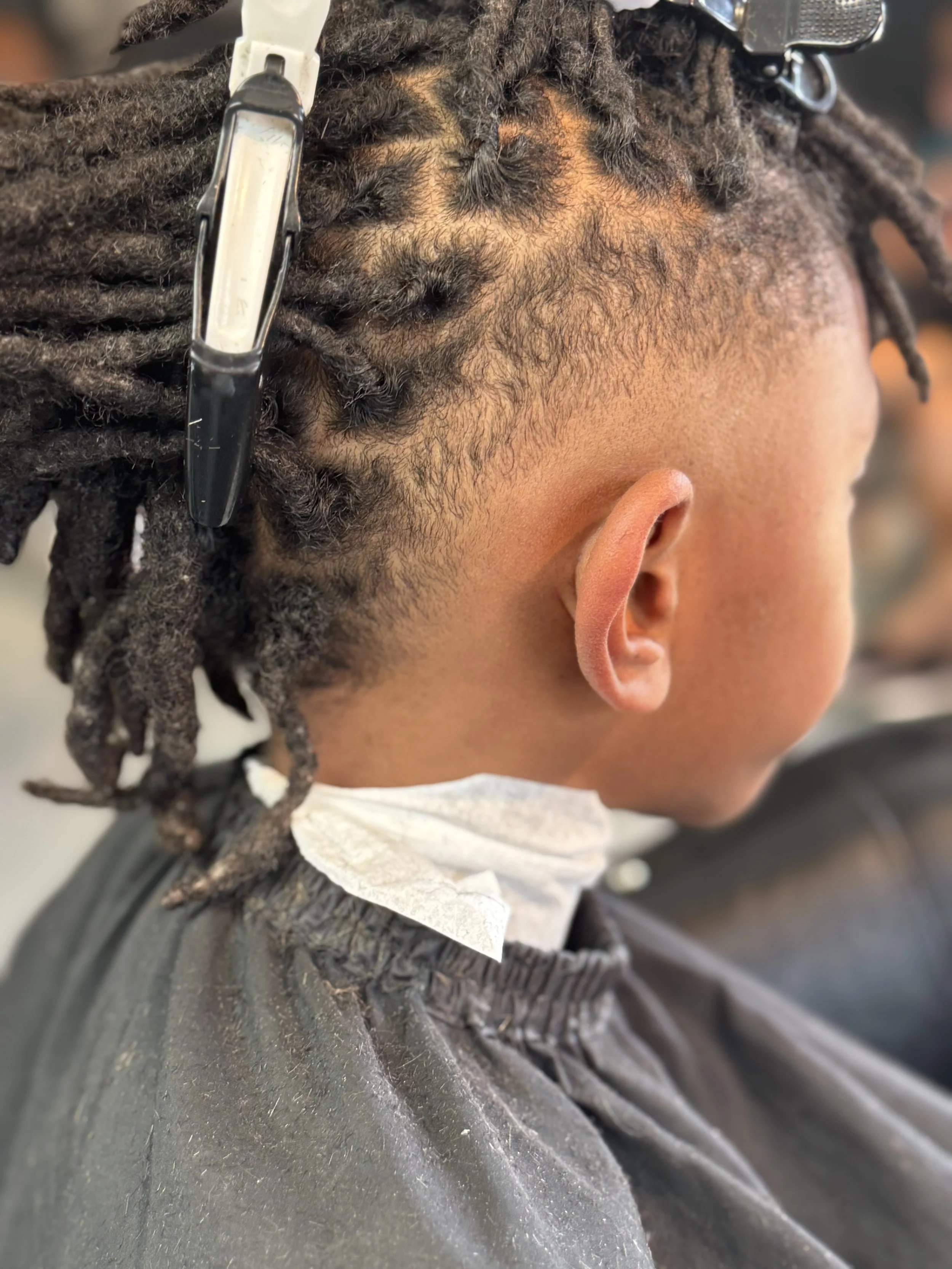 A person with dreadlocks getting a haircut, showing the side profile of their head with freshly trimmed hair, wearing a black haircut cape, a disposable face mask pulled down under the chin, and a clip or razor in the dreadlocks.