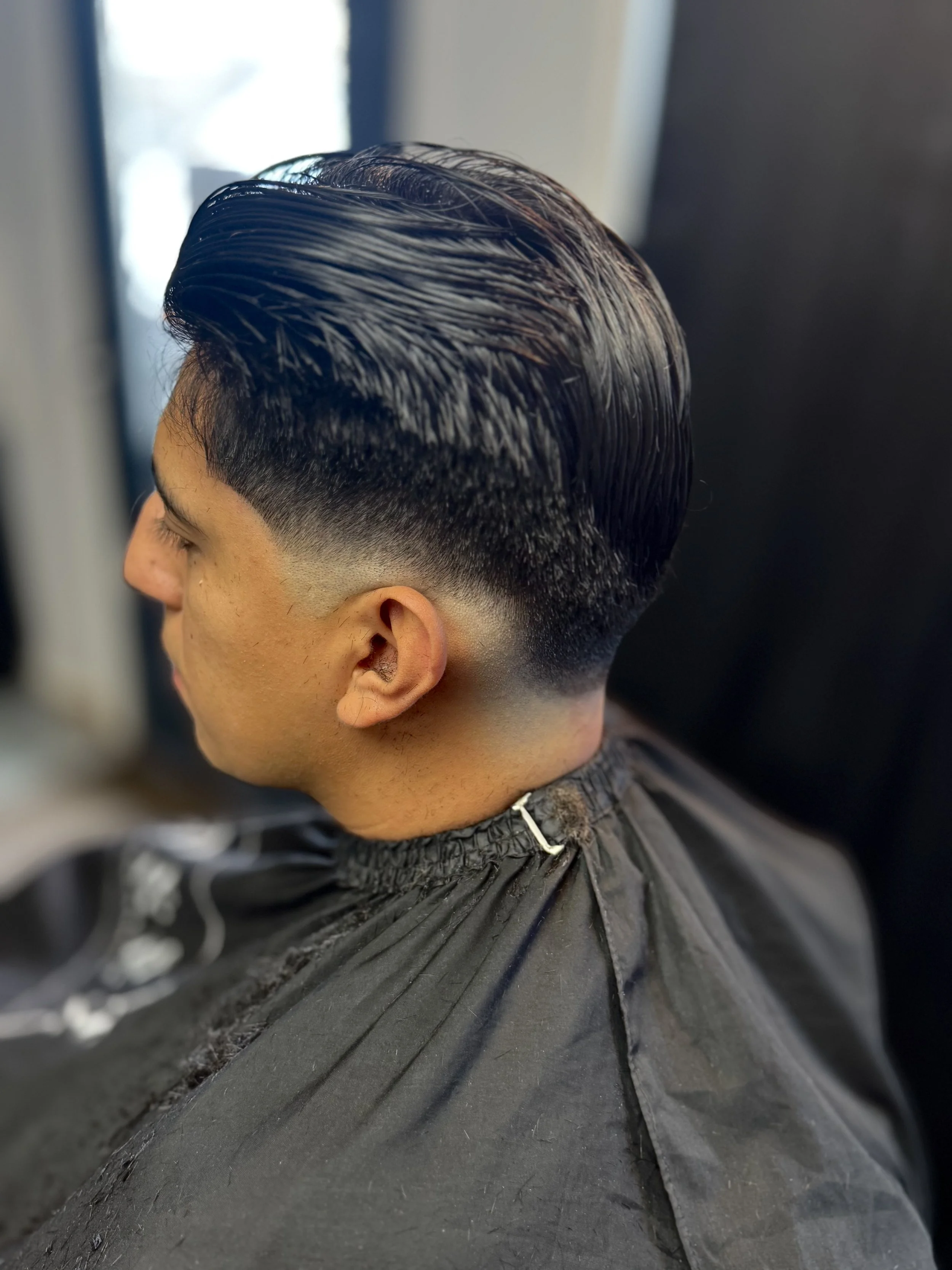 Side view of a man with a fresh, slicked-back hairstyle, featuring a fade haircut, sitting in a barber's chair.