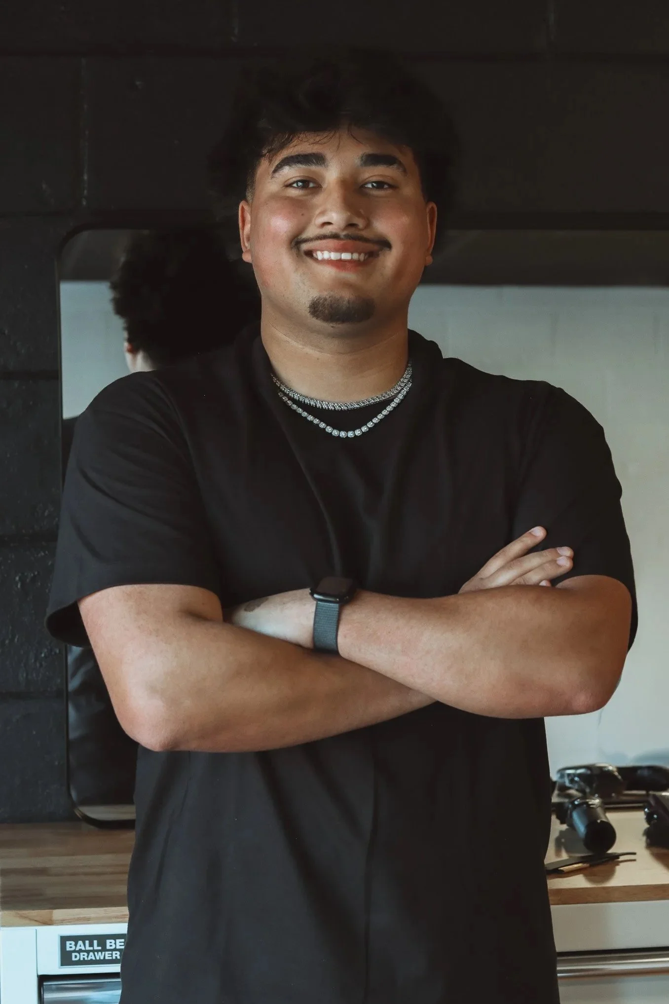 A young man with dark hair, smiling, standing with arms crossed in front of a mirror. He is wearing a black t-shirt, jewelry, and a wristwatch. Behind him, there is a kitchen with a counter, some gadgets, and a drawer labeled 'BALL BE '. a mirror reflects part of his back.