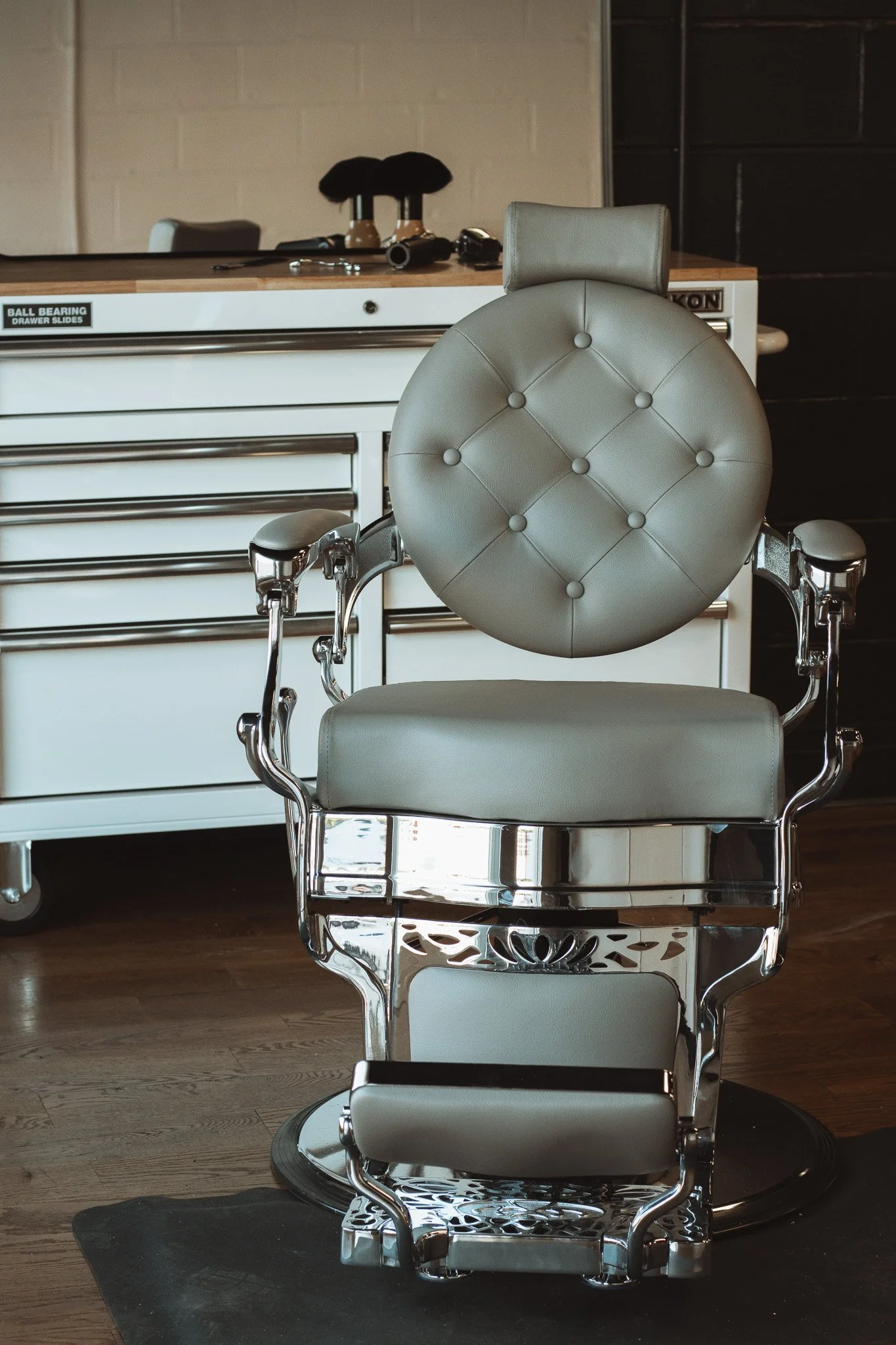A vintage barber chair with beige padding, metal frame, and recline features, situated in a barbershop or salon setting.