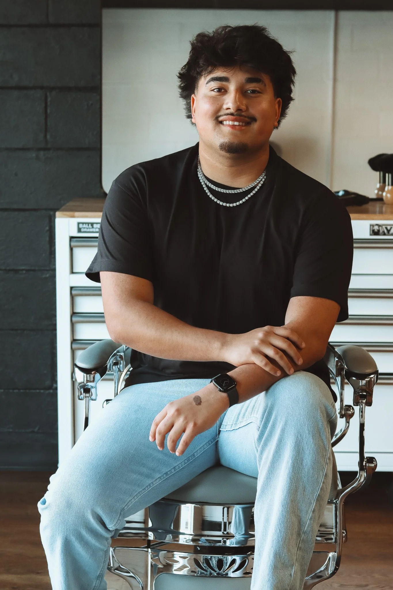 A young man with curly dark hair and a goatee, smiling, sitting on a chair indoors. He is wearing a black t-shirt, light blue jeans, a smartwatch, and a necklace with multiple chains. Behind him are a dark wall, a white cabinet, and some items on top.