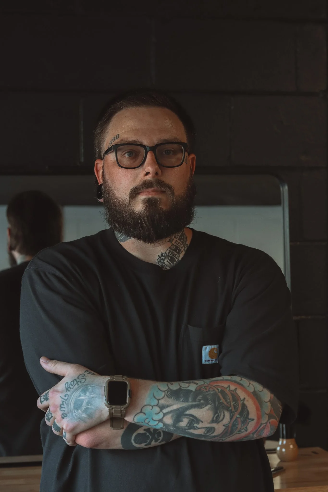 A man with glasses, tattoos, a beard, and a black shirt, standing with arms crossed in front of a mirror in a dark room.