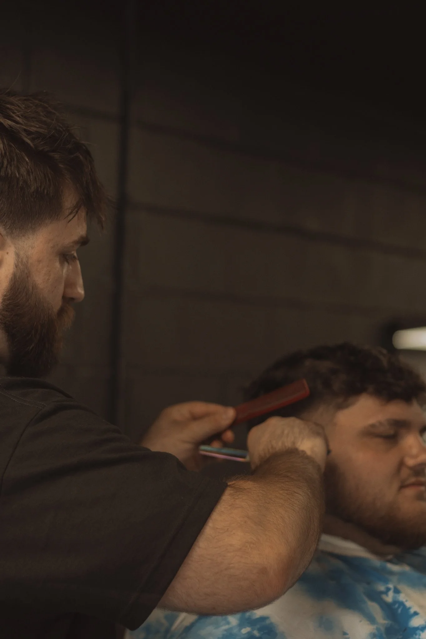 A barber giving a haircut to a young man in a salon with dark walls.