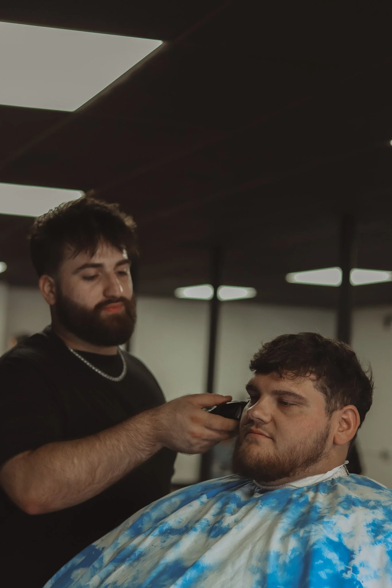 A man getting a haircut in a barbershop, with a barber using clippers on his beard, seated in a barber cape.