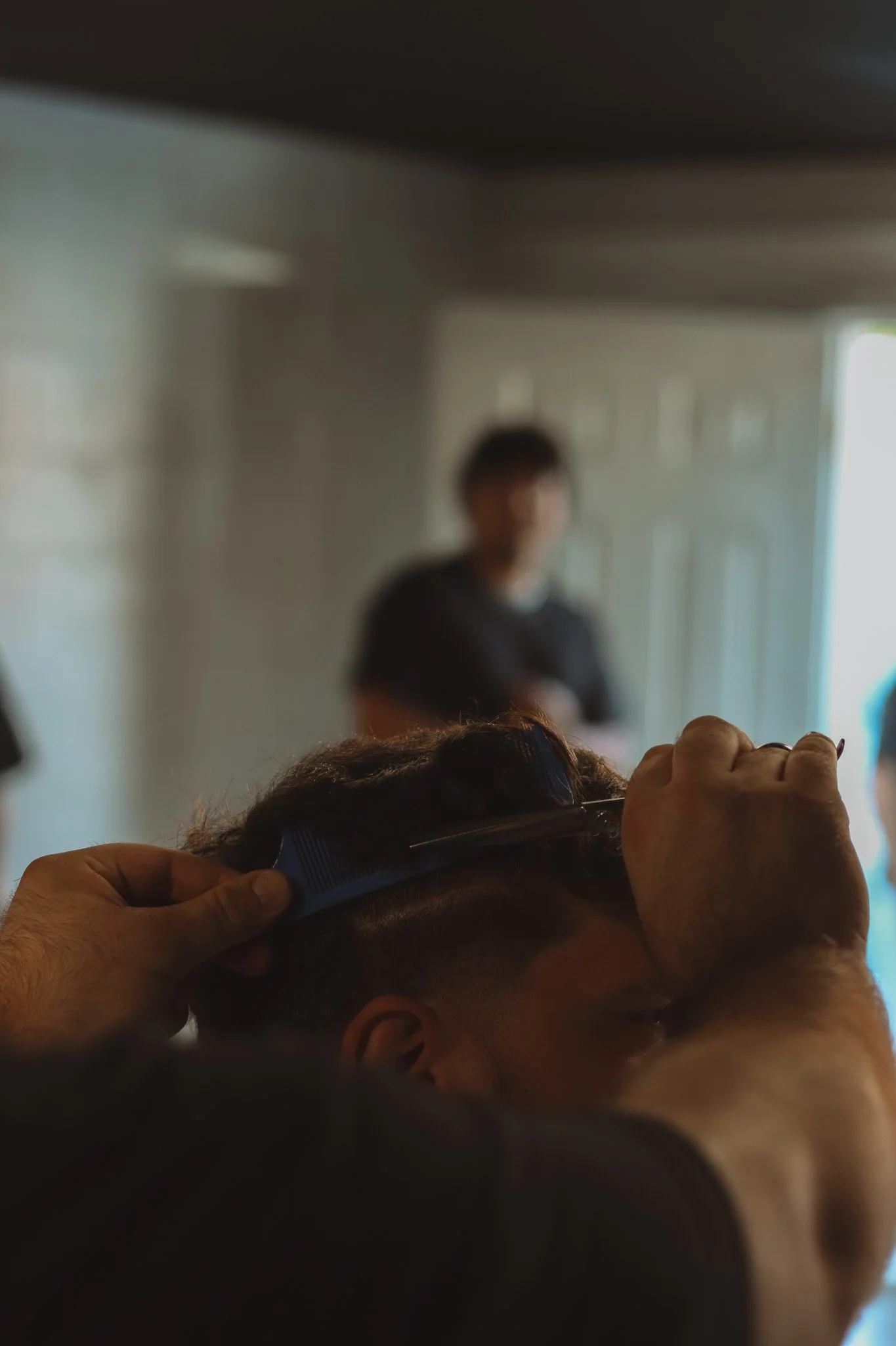 A person is getting a haircut as another person combs their hair with a blue comb, with a blurred person standing in the background near a door.