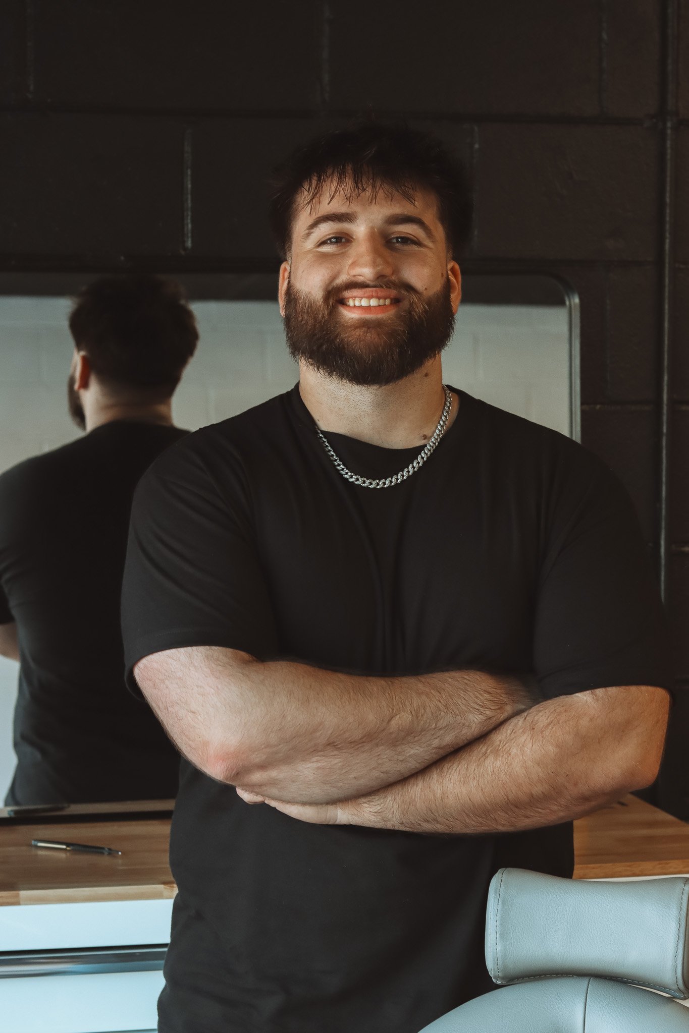 A man with a beard and dark hair, smiling with arms crossed, standing in front of a mirror, wearing a black t-shirt and a silver chain, in a modern room.