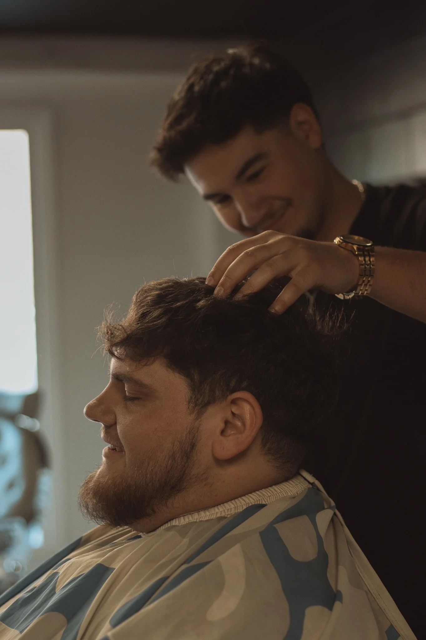 A man getting a haircut at a barbershop, with another man smiling as he trims his hair.