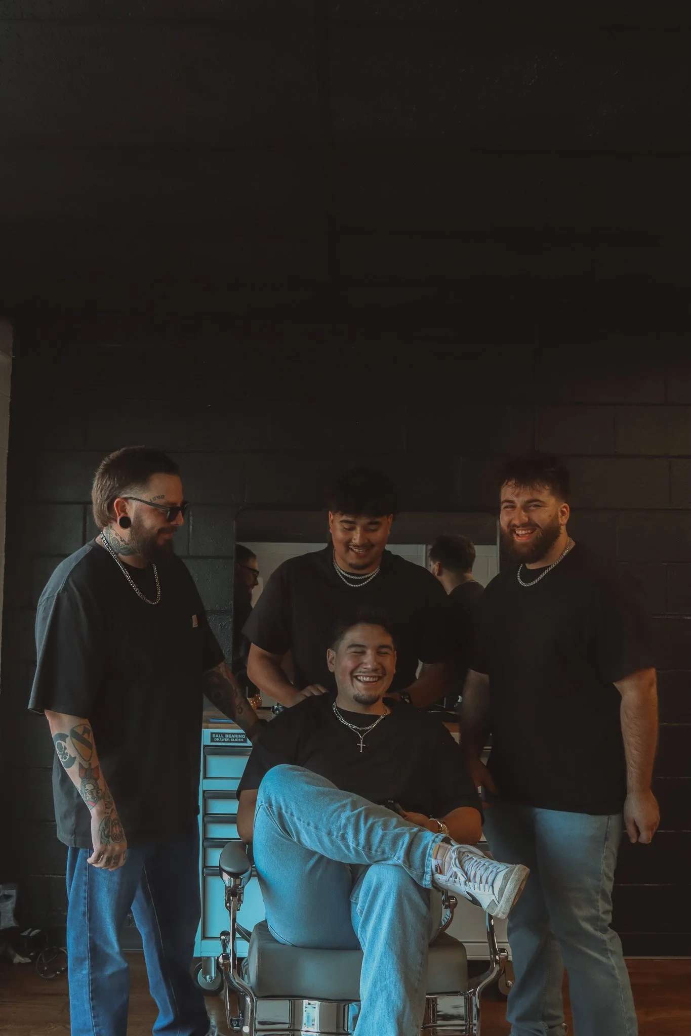 Four smiling men, one sitting in a barber chair and three standing around him, in a barbershop. They are wearing black t-shirts and have tattoos and jewelry. The man in the chair has his legs crossed and is showing a cross necklace.