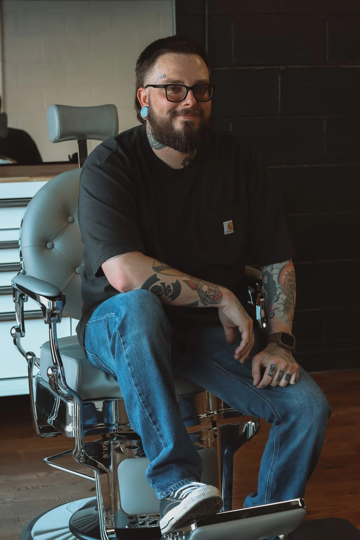 A man with tattoos, glasses, and a beard sitting on an barber chair inside a barbershop, wearing a black t-shirt and jeans.