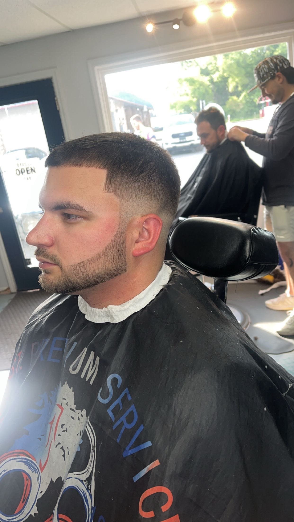 A man receives a fresh haircut at a barbershop. The haircut features a fade with straight lines shaved into the sides. In the background, another person is getting a haircut, and a stylist is working on their hair.