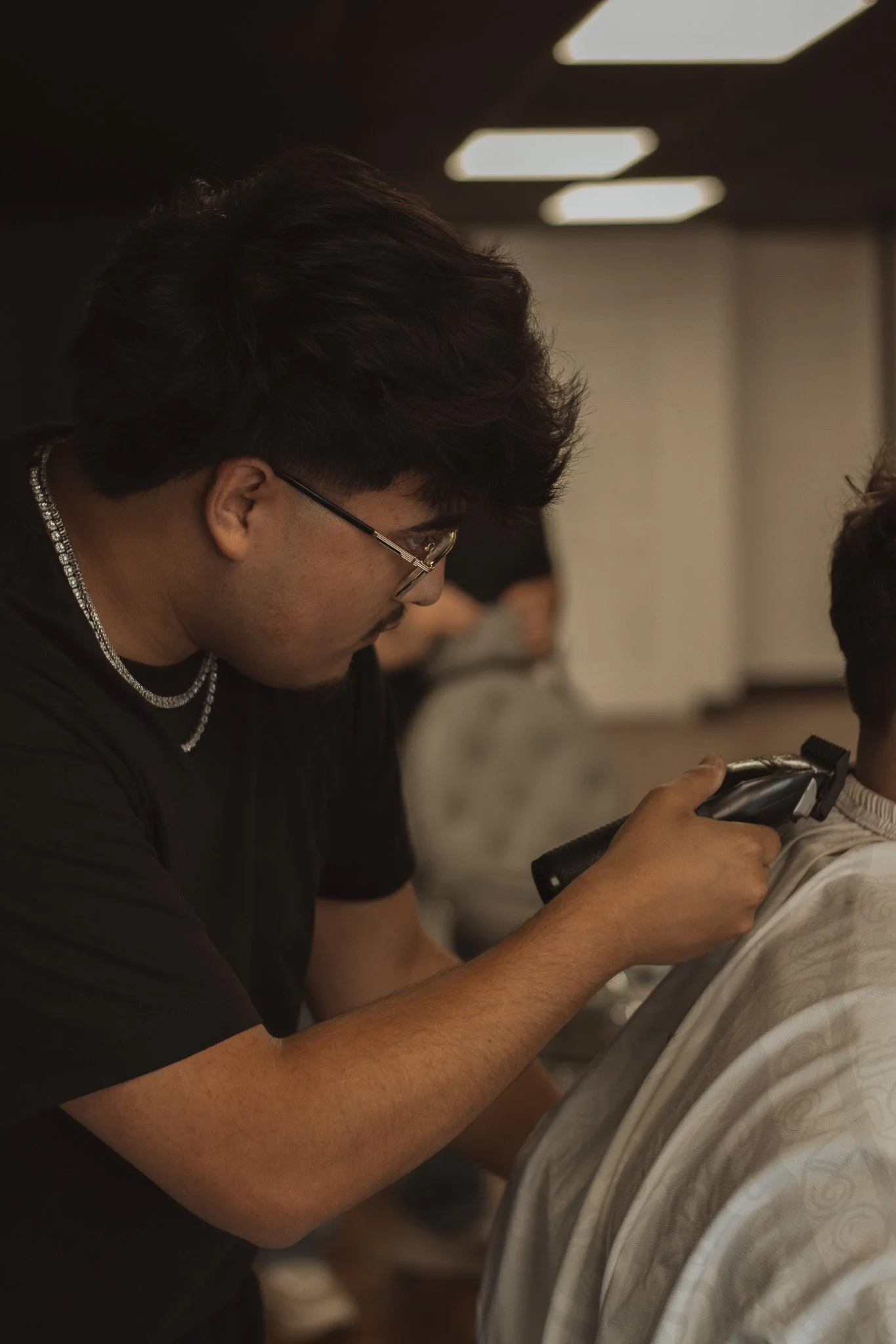 A person with dark hair and glasses is using a straight razor to shave someone else at a barbershop or salon.