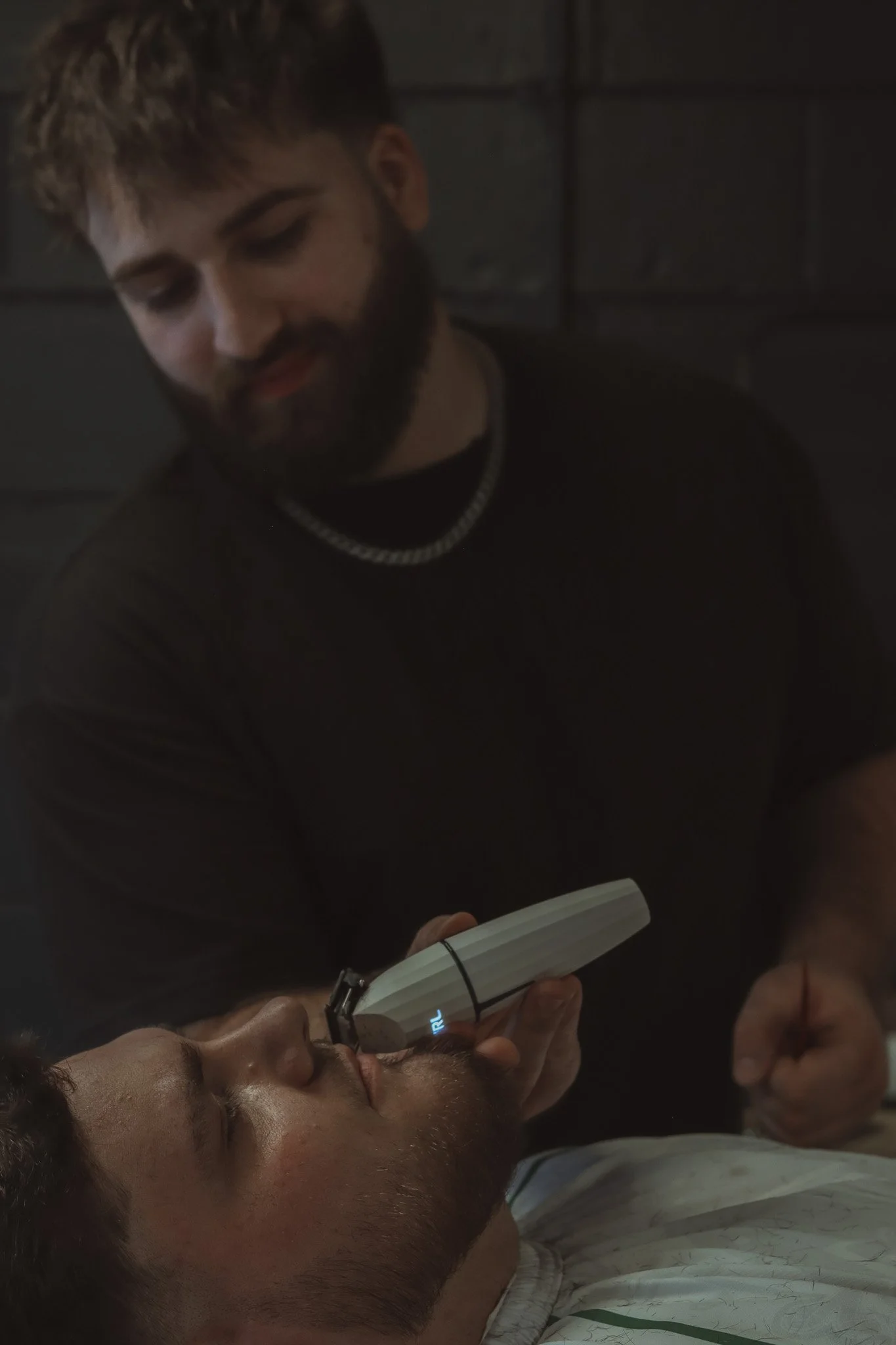 A person lying down with eyes closed, receiving a hair or scalp treatment with a handheld device, while another person attends to them in a dimly lit room.