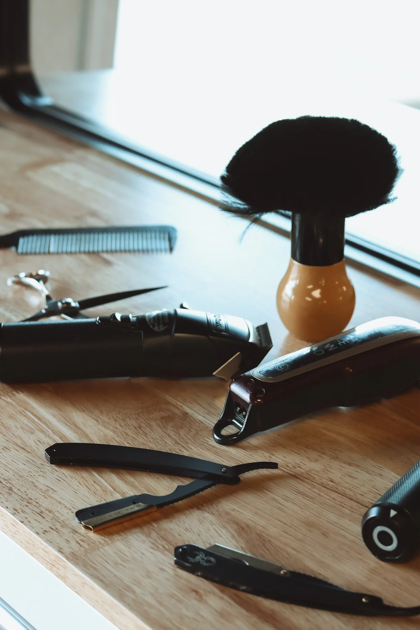Collection of barber tools on a wooden surface, including a hairbrush, hair clippers, a straight razor, a shaving razor, a spray bottle, and a styling brush.