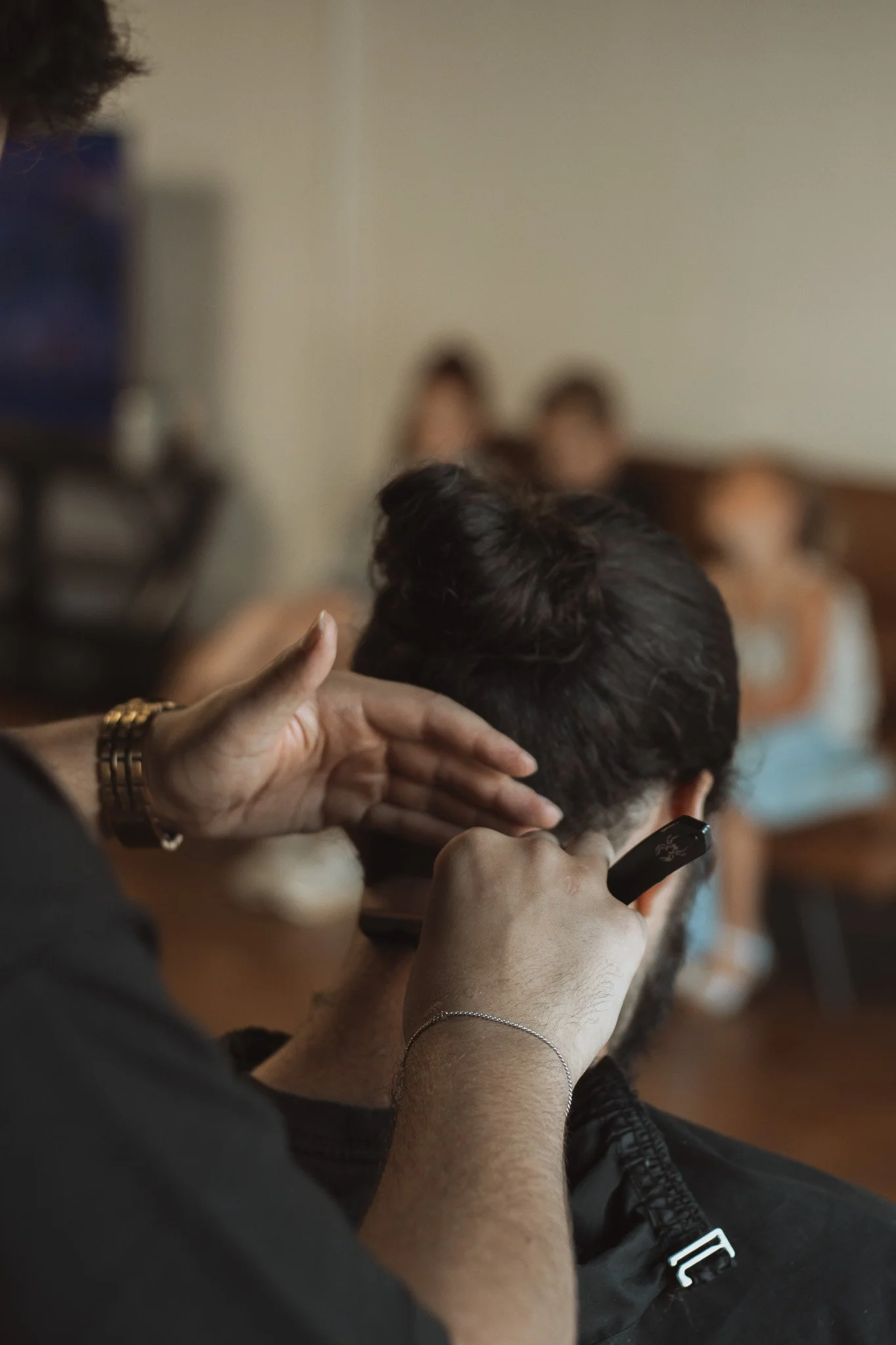 Person receiving a haircut with scissors, partially visible, while a barber trims the man's hair in a salon or barber shop setting with people seated in the background.