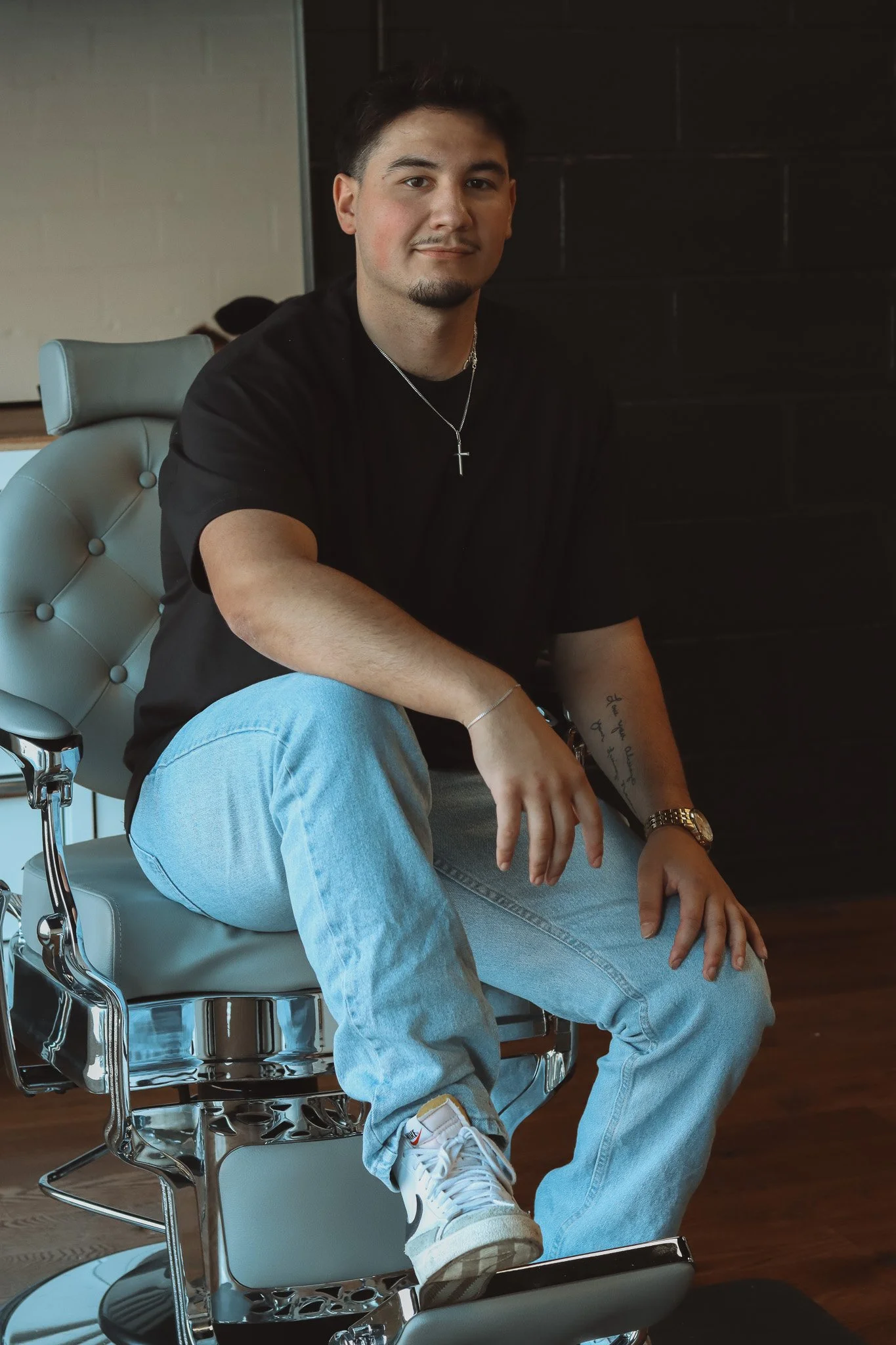 A young man with dark hair and a goatee sits on a barber chair, wearing a black t-shirt, light jeans, white sneakers, a silver watch, and a cross necklace.