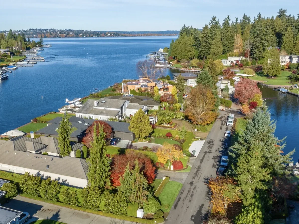 Aerial view of houses and boats along a calm river with lush green and fall-colored trees, and a forested hillside in the background.