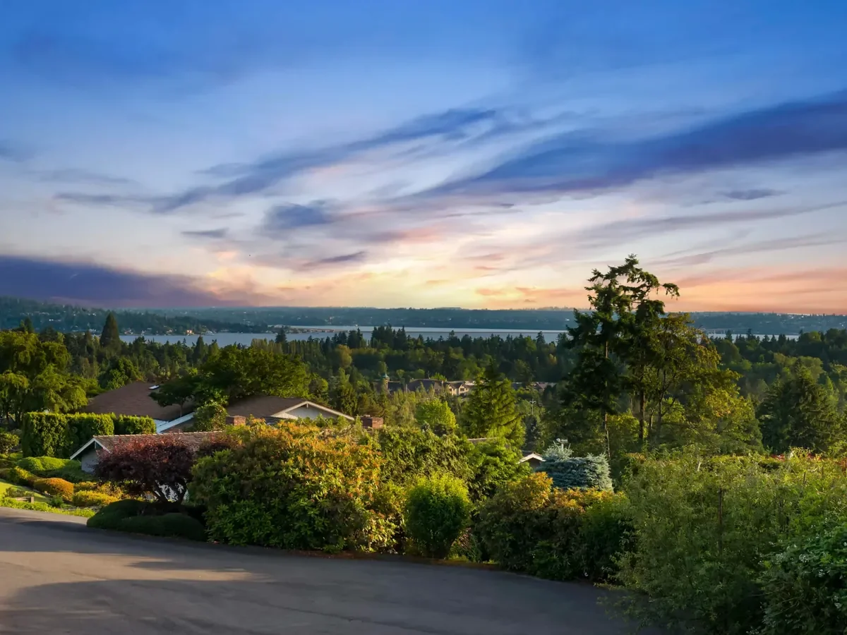 Scenic view of a suburban neighborhood at sunset with houses, lush green trees, and a body of water in the distance under a colorful sky.
