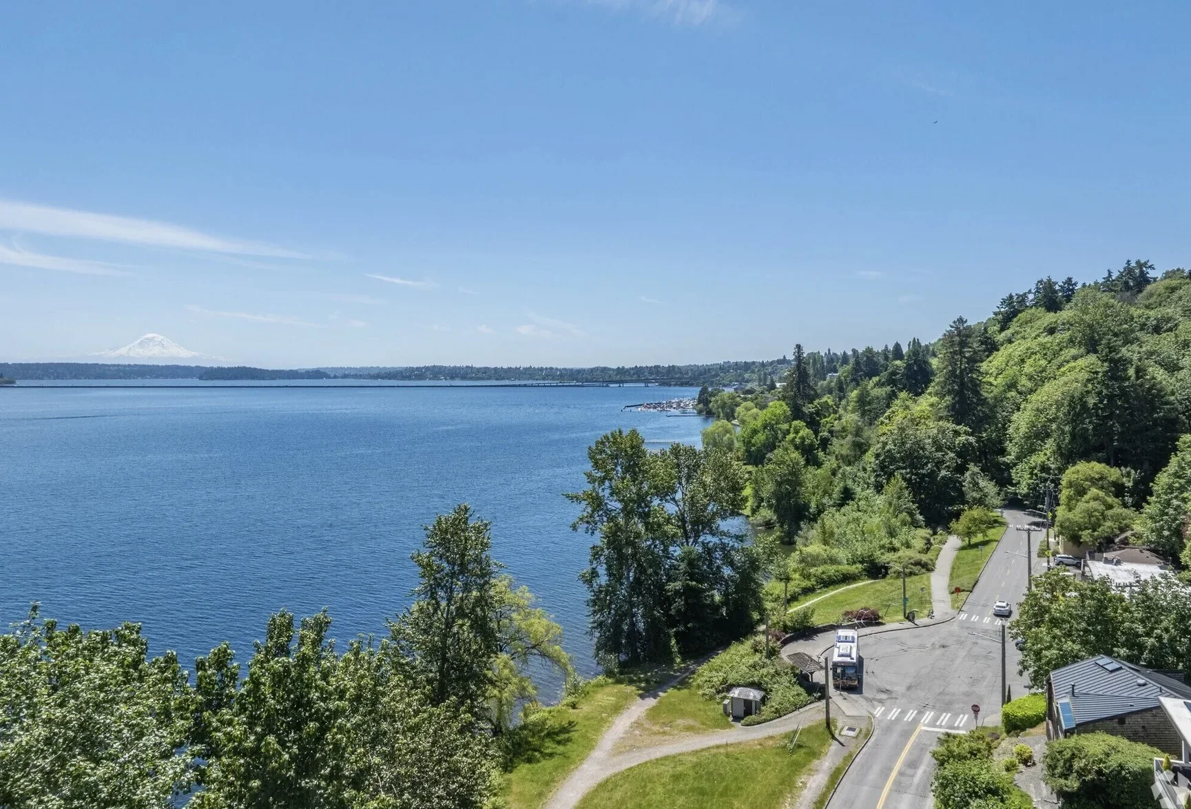 A scenic view of a large lake with clear blue water, surrounded by lush green trees and hills. A road with a few cars runs along the shoreline, and in the distance, a snow-capped mountain peaks above the horizon on a bright, sunny day.