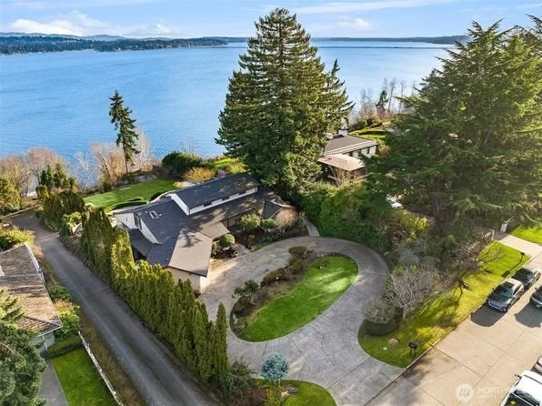 Aerial view of a residential lakeside property with a curved driveway, green lawn, tall trees, neighboring houses, and a large body of water in the background.